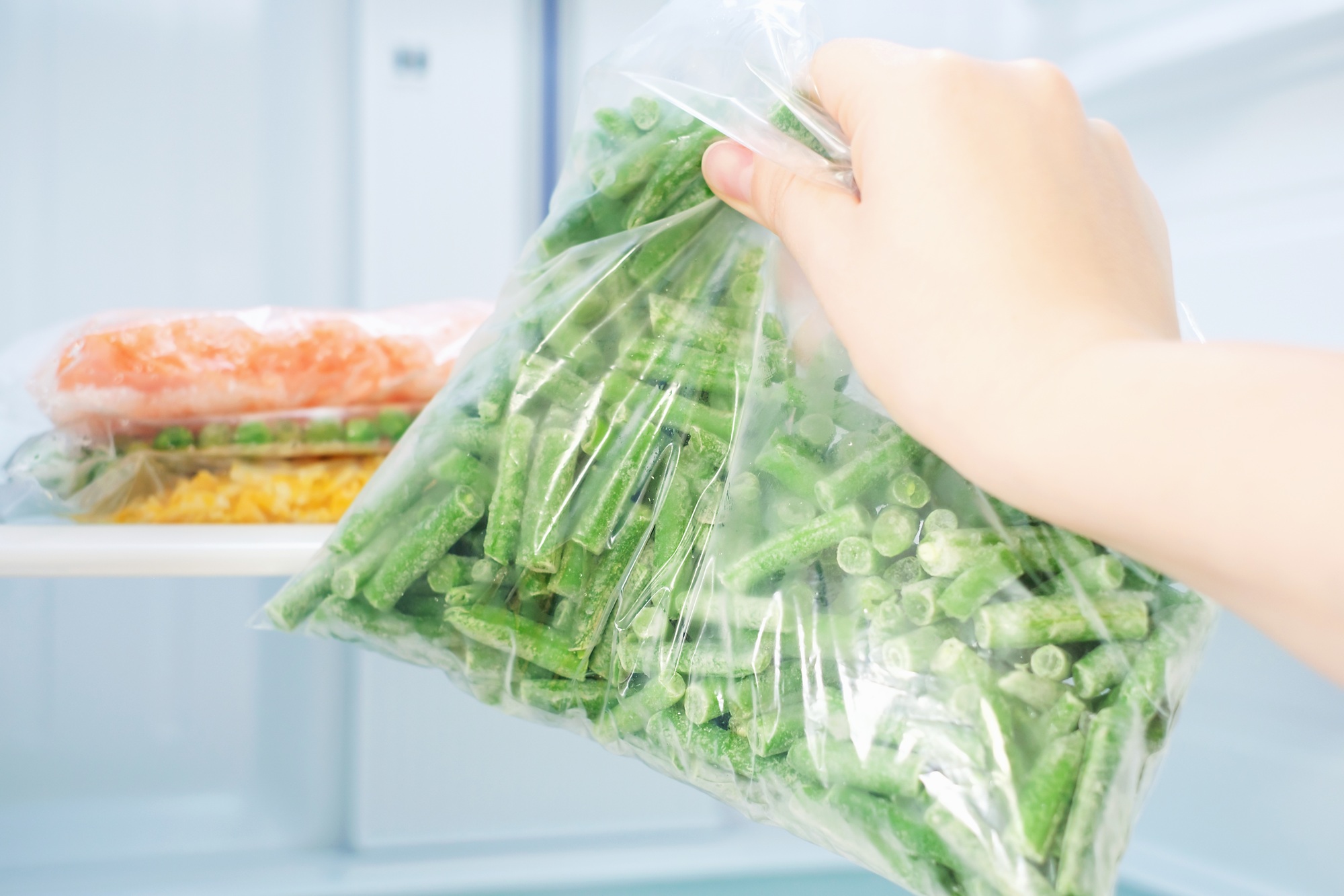 Female hand holds a plastic bag with frozen green beans on the background of freezer.