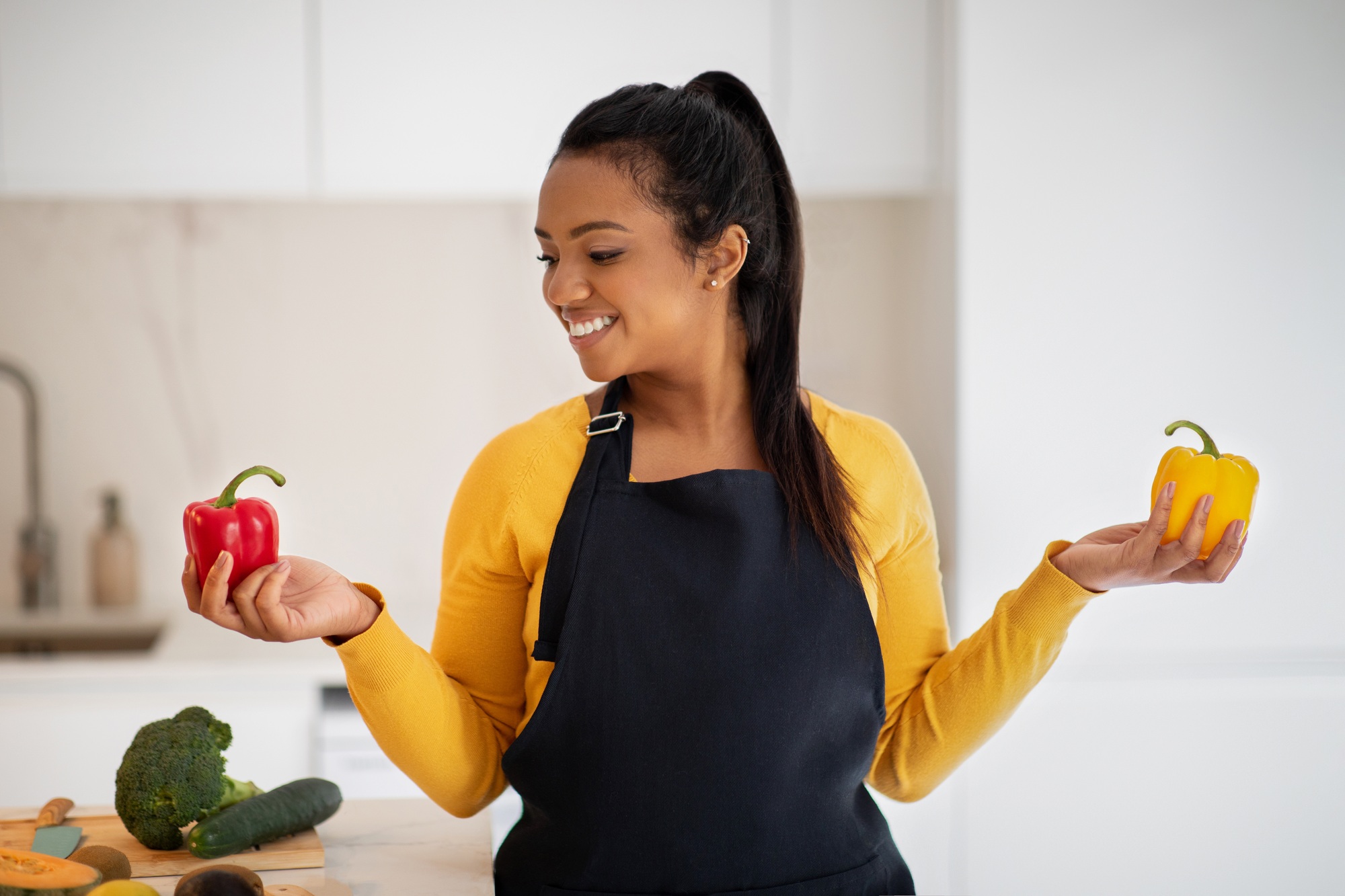 Cheerful millennial african american female in apron chooses peppers in hands at table with