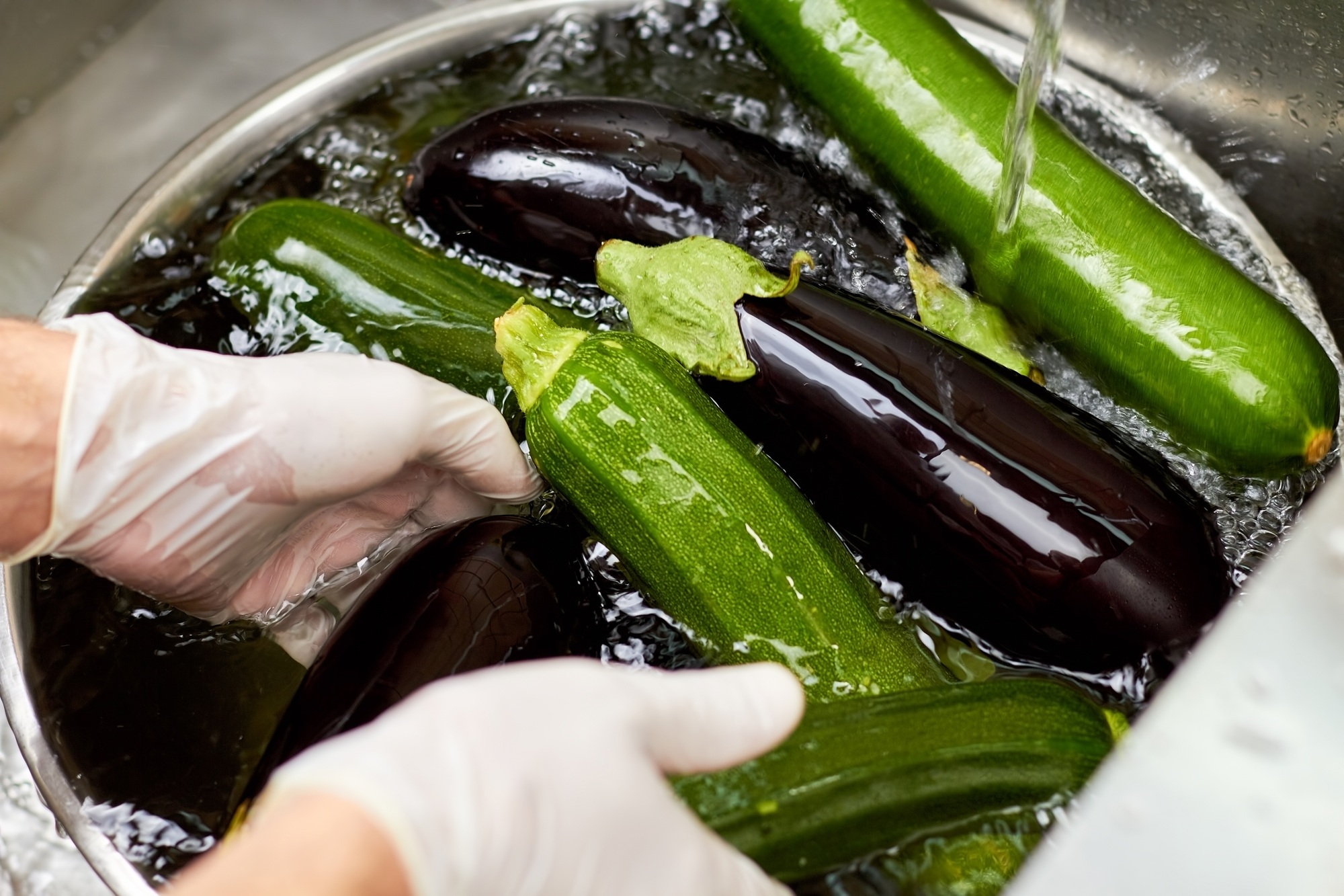 Washing heap of zucchinis and eggplants in metal bowl.