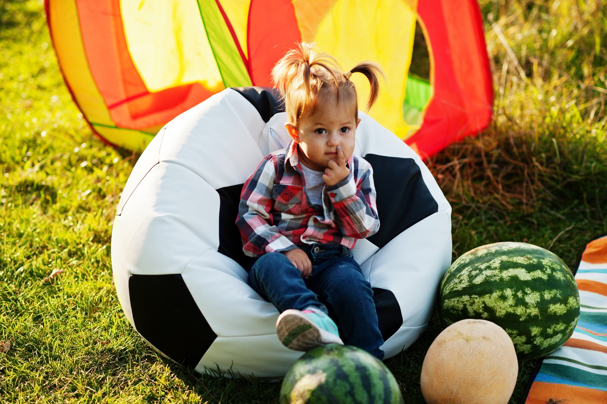 Cute baby girl in checkered shirt sit on football ball pouf with watermelons.