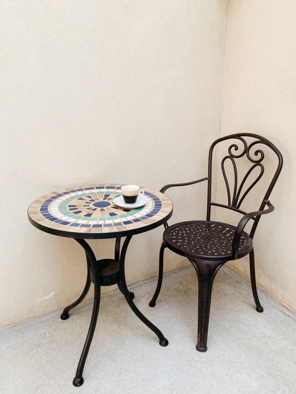 Photo of a small mosaic-topped table next to a wrought iron chair, corner angle, patio corner setting, bright natural light, containing a small espresso cup on the table, iPhone photo quality.