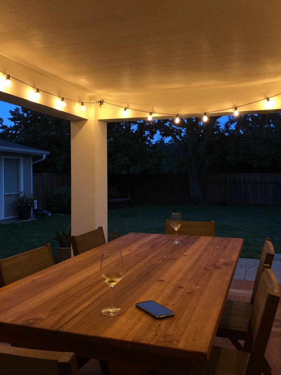 Photo of warm string lights draped above a wooden dining table, wide room view, evening backyard setting, soft indoor lighting from bulbs, containing a half-full wine glass on the table, iPhone photo quality.