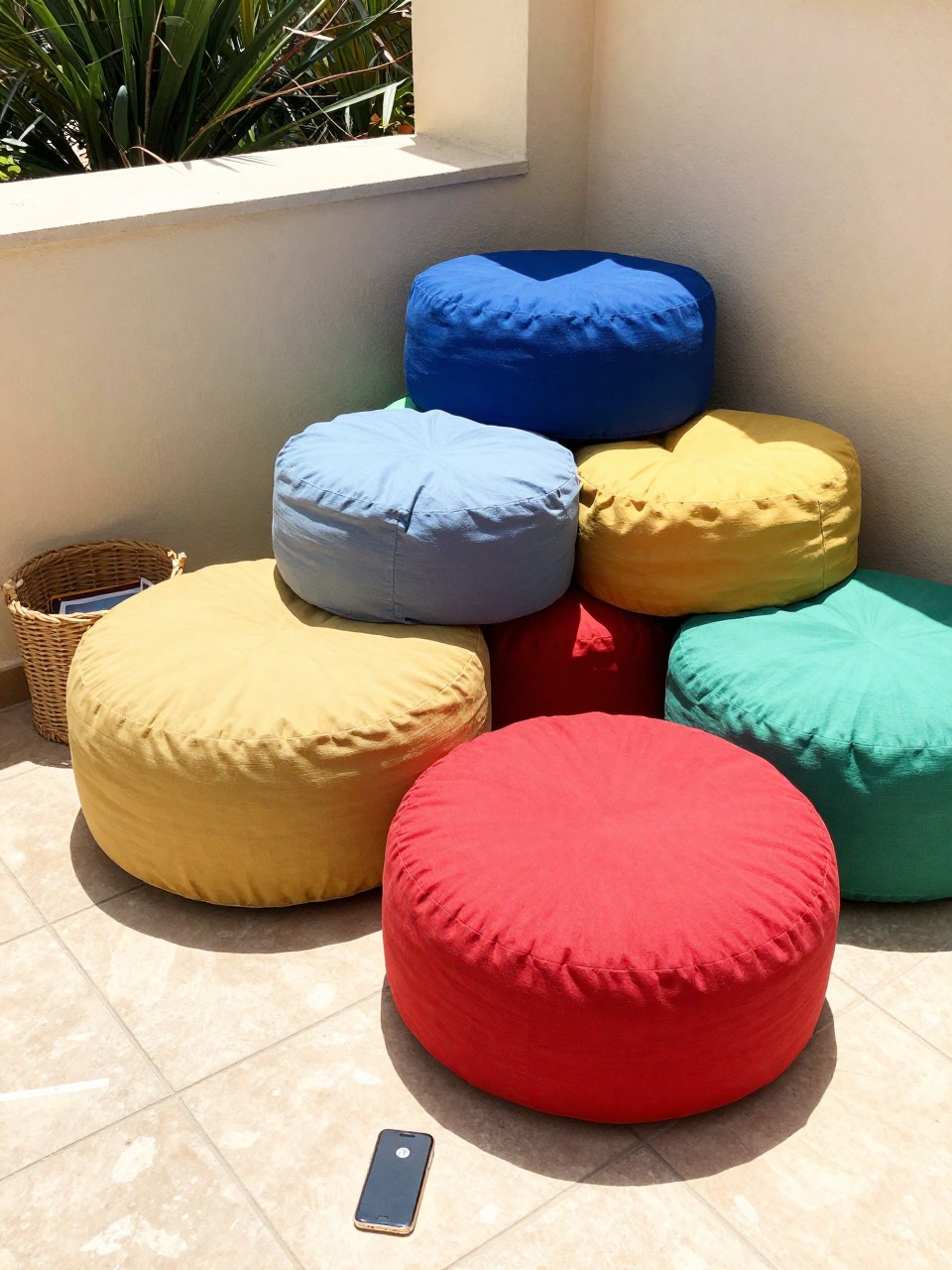 Photo of a patio corner with a colorful pile of mismatched poufs, corner angle view, setting on a sunny patio with tile floors, bright midday light, containing a woven magazine holder nearby, iPhone photo quality.
