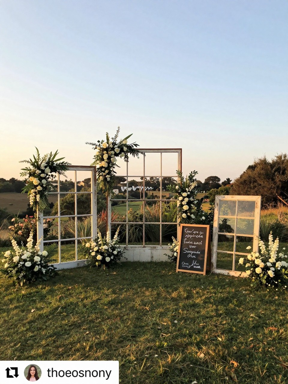 Photo of a outdoor wedding setup with large old window frames as a backdrop decorated with flowers, wide view, setting in a garden wedding, golden hour, containing a chalkboard sign on one frame with the couple's names, iPhone photo quality.