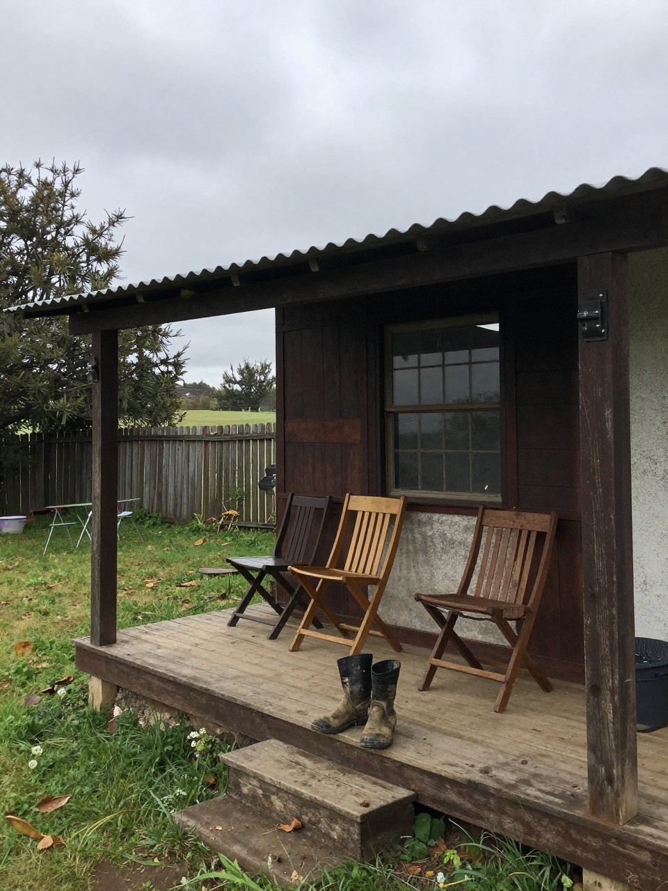 Photo of a small tin roof overhang covering chairs in a rustic country backyard, corner angle, overcast day, containing a pair of muddy boots left by the steps, iPhone photo quality.