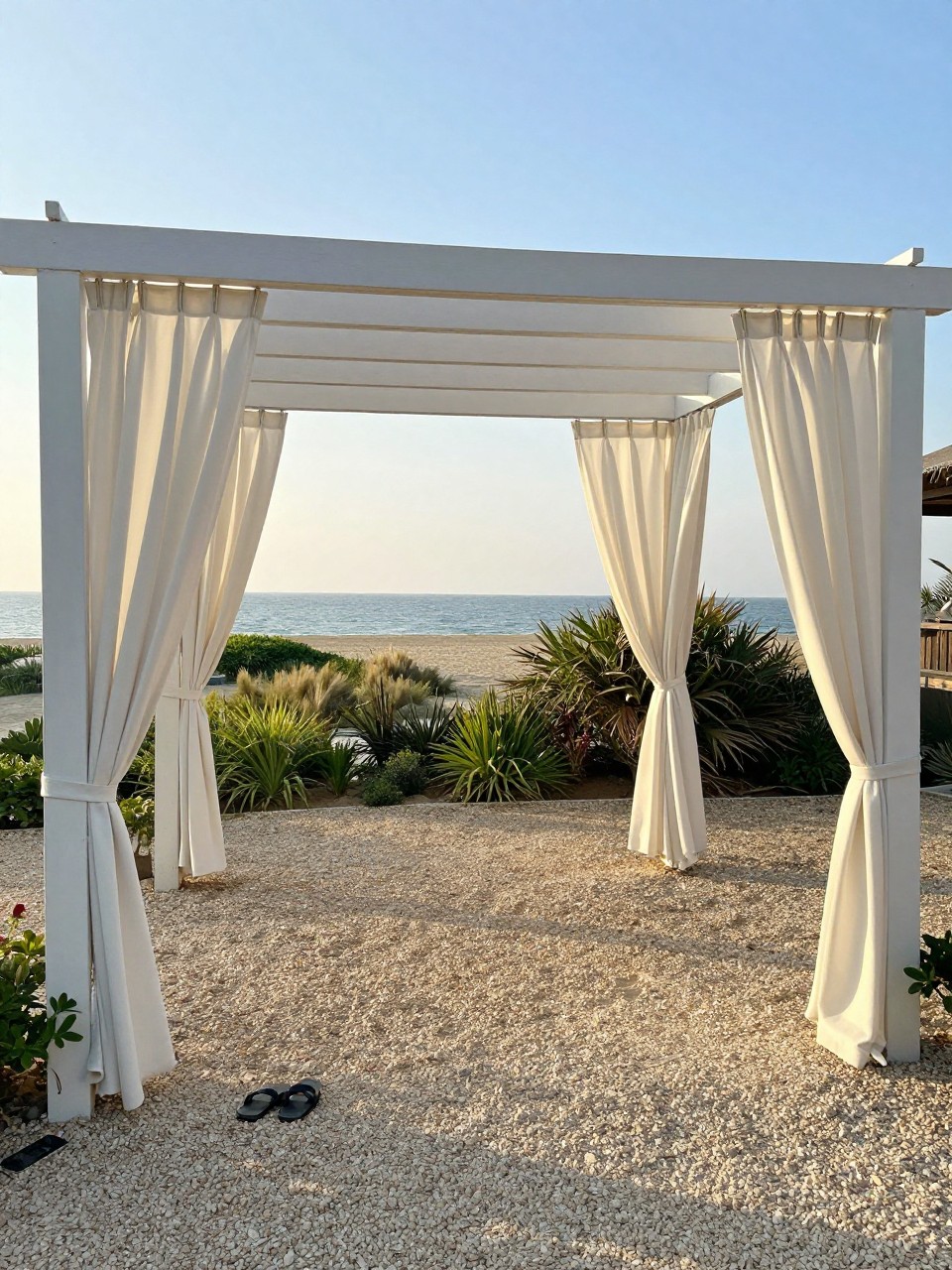 Photo of a pergola with white outdoor curtains tied back at the corners, wide view, setting in a beachy garden with sand-colored gravel, late afternoon light, containing a pair of sandals on the ground, iPhone photo quality.