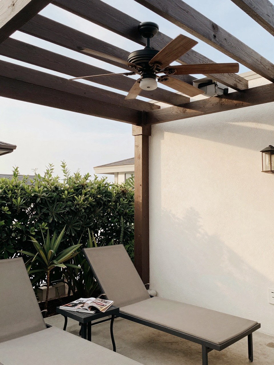 Photo of a pergola with a wooden ceiling fan spinning overhead, corner angle view, setting in a covered patio with lounge chairs, soft afternoon light, containing a magazine on a side table, iPhone photo quality.