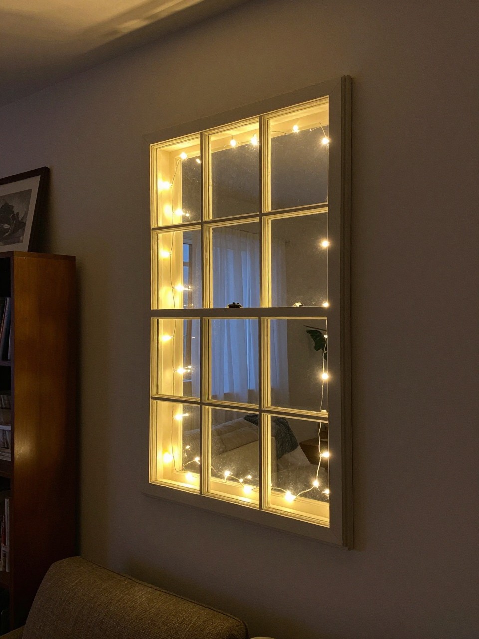 Photo of a old window frame hung on a wall with warm string lights glowing through the panes, corner angle view, setting in a cozy living room at night, soft ambient light, containing a bookshelf nearby, iPhone photo quality.