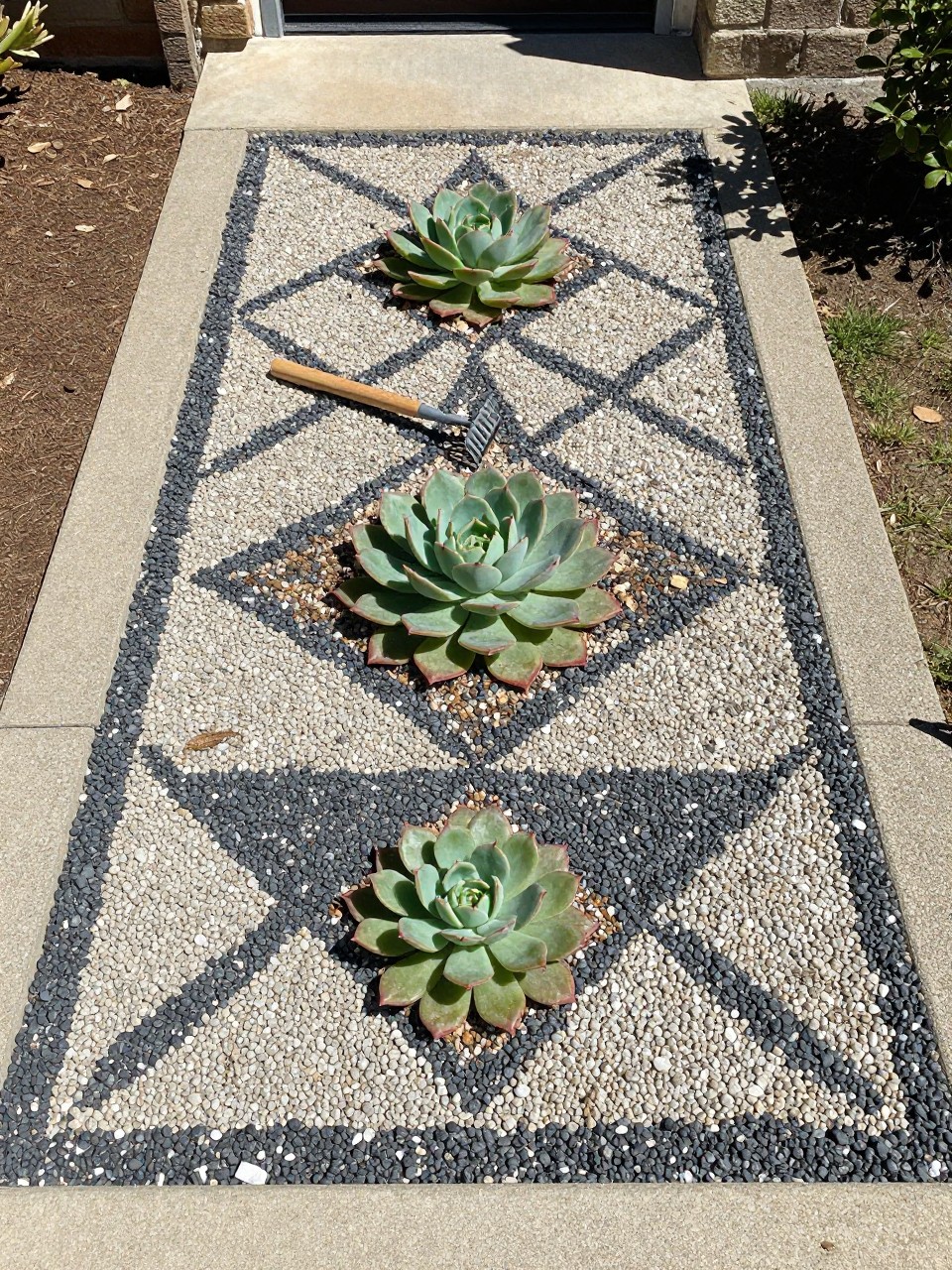 Photo of a geometric succulent mosaic pattern in a gravel bed, straight-on overhead view, setting in a front entry garden, bright midday light, containing a small hand rake nearby, iPhone photo quality.