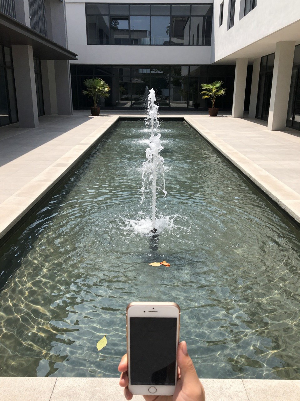 Photo of a rectangular reflecting pool with a single water jet in the center, straight-on view, setting in a courtyard with modern architecture, bright midday light, containing a single leaf floating on the surface, iPhone photo quality.