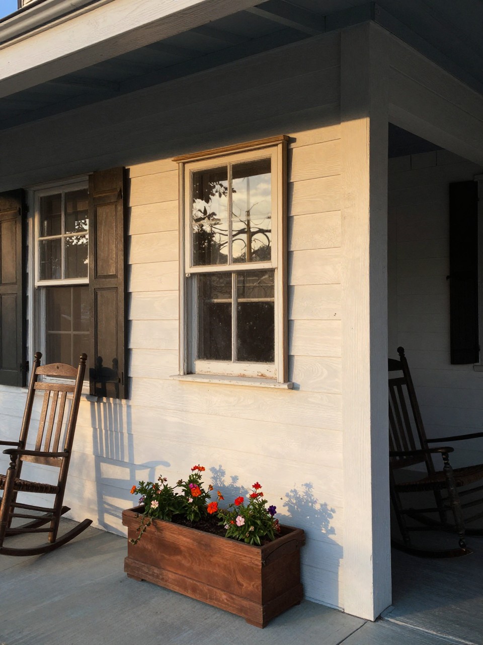 Photo of a covered porch with an old window frame hanging on the wall, a small planter box at the bottom with trailing flowers, corner angle view, setting in a farmhouse porch, late afternoon light, containing a rocking chair nearby, iPhone photo quality.
