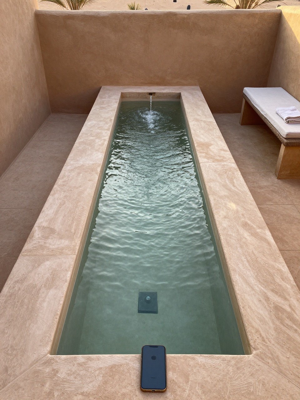 Photo of a long rectangular water feature with a gentle trickle, straight-on view, setting in a shaded desert patio corner, golden hour, containing a folded linen towel on a nearby bench, iPhone photo quality.