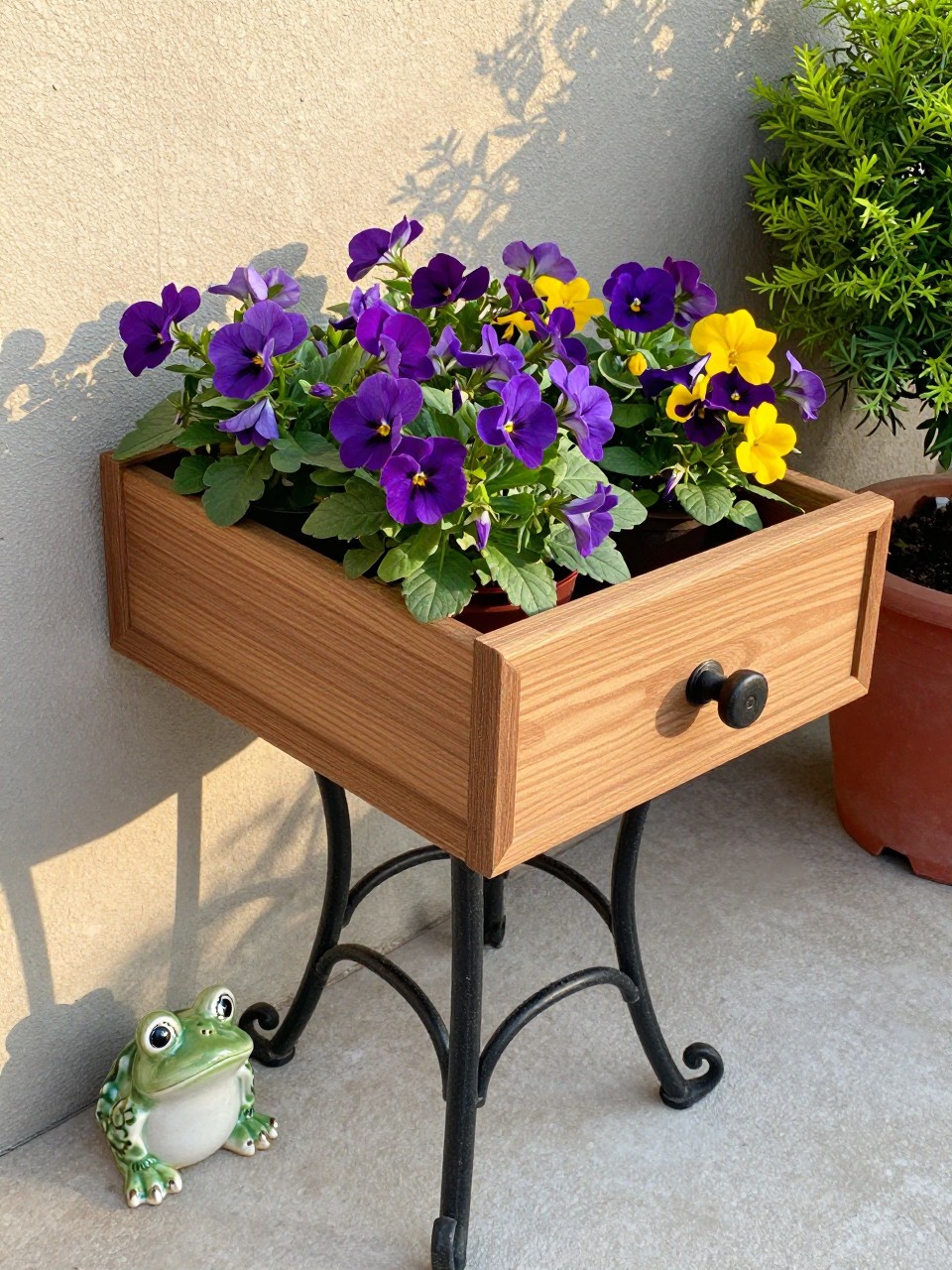 Photo of a wooden drawer sitting on a wrought iron plant stand, filled with purple and yellow flowers, corner angle view, setting on a patio, late afternoon light, containing a small ceramic frog near the stand, iPhone photo quality.