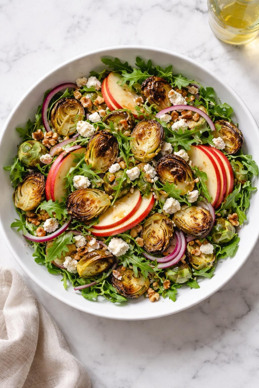 Image Prompt: Realistic top-down editorial food photography of a roasted Brussels sprout salad in a white serving bowl on a clean marble countertop, roasted Brussels sprouts with thin apple slices, chopped walnuts, arugula, red onion, and goat cheese, light apple cider vinaigrette over the top, bright natural light, soft shadows, clean editorial plating, realistic textures, no people, no hands, no text, no watermarks, no props with writing.