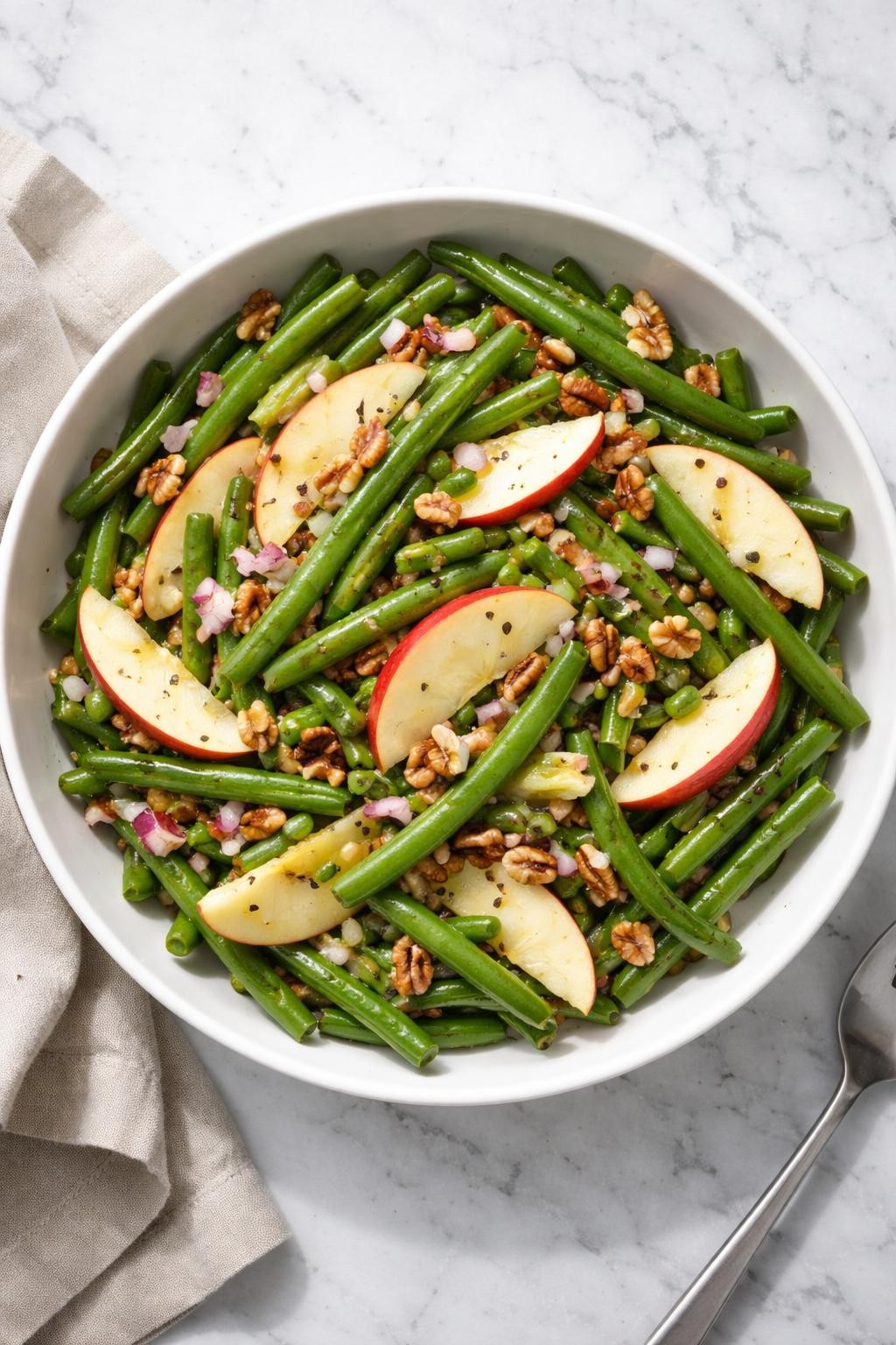 Image Prompt: Realistic top-down editorial food photography of a cold green bean salad with apples and pecans in a white serving bowl on a clean marble countertop, chilled green beans tossed with thin apple slices, chopped pecans, red onion, and light apple cider vinaigrette, bright natural window lighting, soft shadows, clean composition, high detail, realistic food texture, no people, no hands, no text, no watermarks, no props with writing.