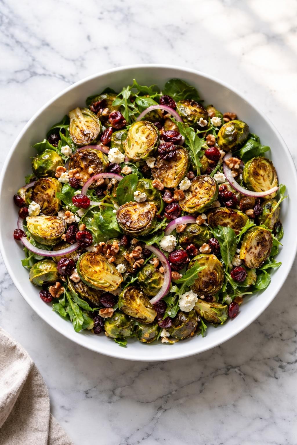 Image Prompt: Realistic top-down editorial food photography of a roasted Brussels sprout salad in a wide white bowl on a clean white and gray marble countertop, caramelized roasted Brussels sprouts tossed with mixed greens, dried cranberries, chopped pecans, thin red onion, and crumbled feta cheese, light balsamic dressing drizzled over the top, bright natural window lighting, soft shadows, clean composition, high detail, realistic food texture, cozy homemade salad mood, no people, no hands, no text, no watermarks, no props with writing.