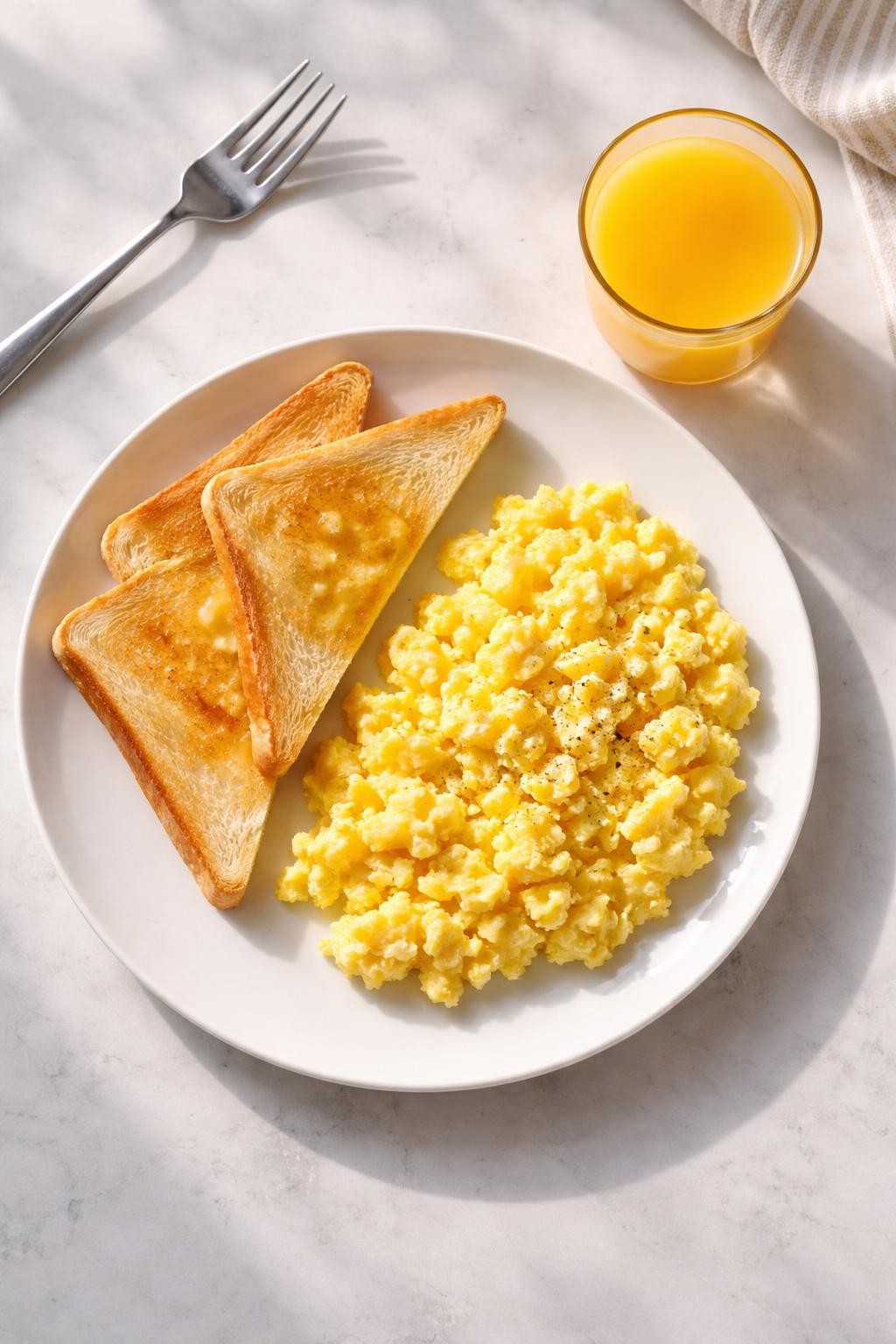"Top-down realistic shot of a white plate with fluffy, smooth scrambled eggs and two slices of buttered white toast cut into triangles. A small glass of orange juice. Bright morning light, simple breakfast-for-dinner, no people, no hands, no text, no watermarks."