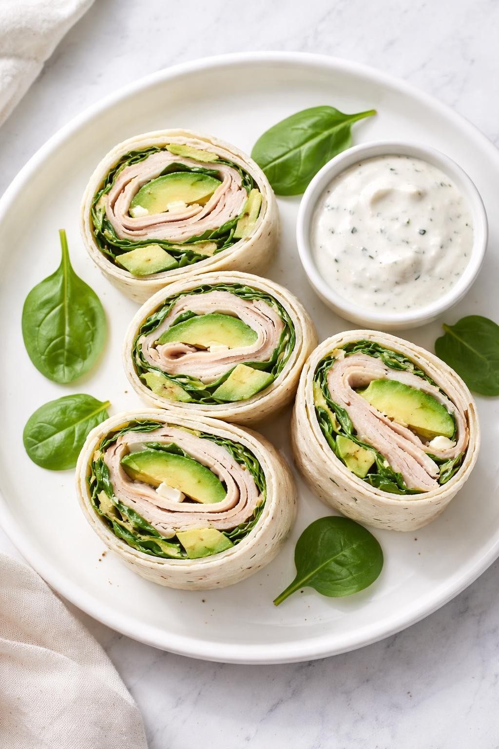 Photo prompt: Realistic top-down food photography of a white plate with four tortilla roll-ups sliced into pinwheels, showing layers of turkey, avocado, spinach, and cream cheese. A small container of ranch dip on the side. Bright natural light, no-sandwich lunch box idea mood, no people, no hands, no text, no watermarks, no props with writing.