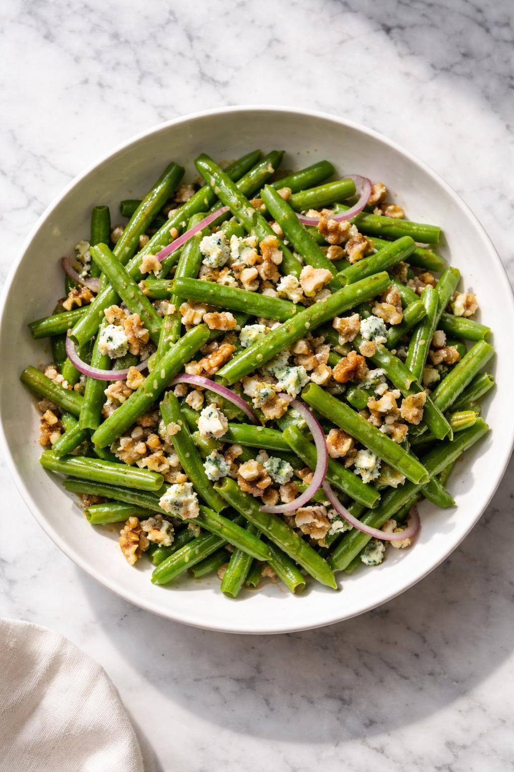 Image Prompt: Realistic top-down editorial food photography of a cold green bean salad with walnuts and blue cheese in a shallow white bowl on a clean white and gray marble countertop, chilled green beans with chopped walnuts, blue cheese crumbles, red onion, and light vinaigrette, bright natural window lighting, soft shadows, elegant homemade salad styling, high detail, realistic food texture, no people, no hands, no text, no watermarks, no props with writing.