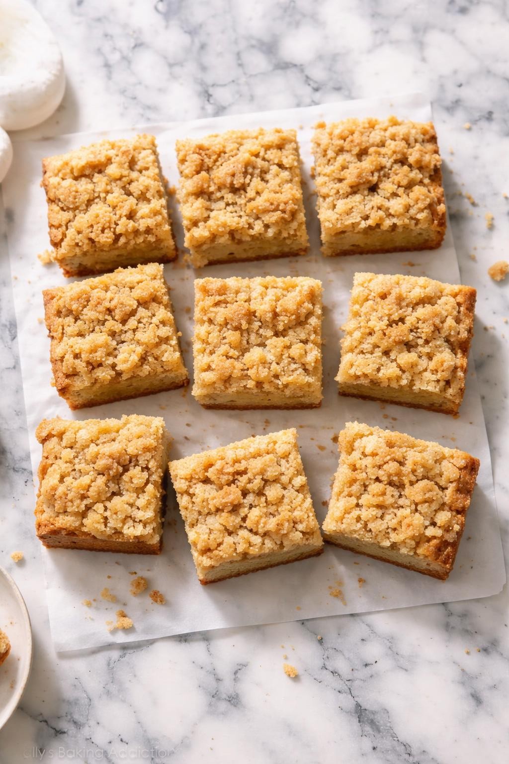 Image Prompt: Realistic top-down editorial food photography of vegan banana crumb cake sliced into squares on a white and gray marble countertop, soft banana cake with a buttery-looking crumb topping, cozy bakery-style mood, bright natural daylight, clean composition, no people, no hands, no text, no watermarks, no props with writing.