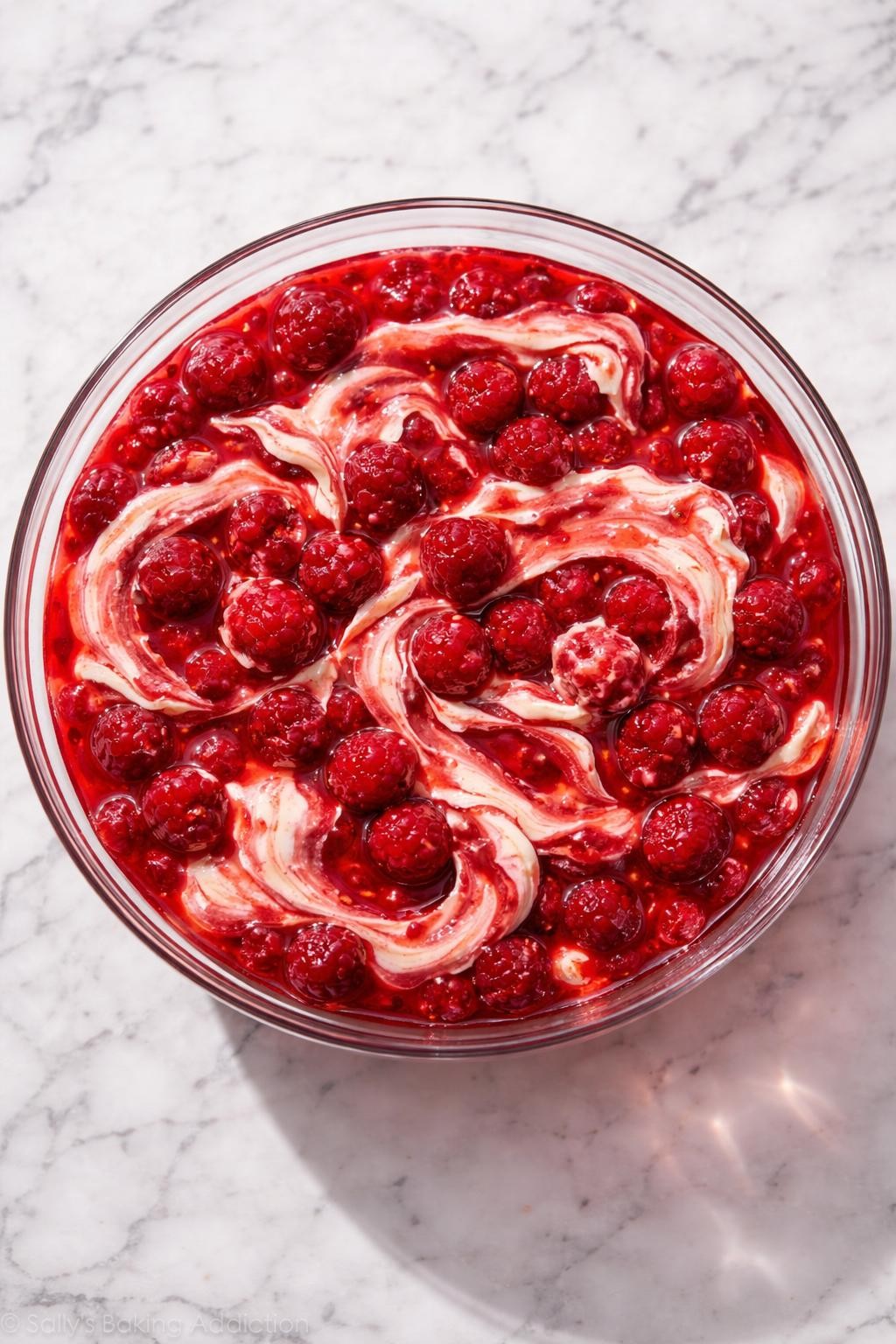 Image Prompt: Realistic top-down editorial food photography of raspberry Jello salad with cream cheese in a clear glass bowl on a clean marble countertop, ruby red gelatin salad with raspberries and creamy swirls of softened cream cheese, chilled retro dessert salad styling, bright natural light, soft shadows, realistic food texture, high detail, no people, no hands, no text, no watermarks, no props with writing.