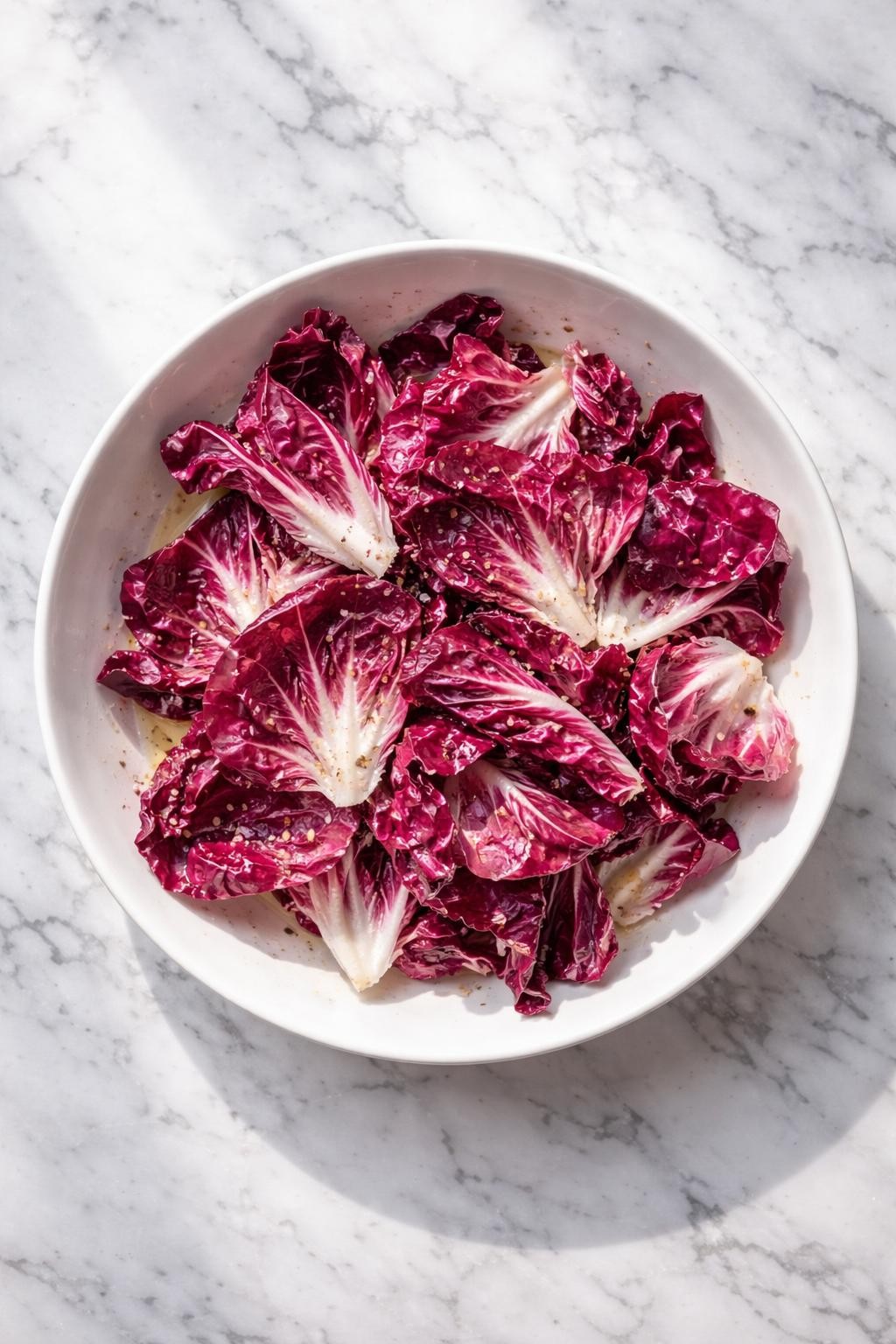 Image Prompt: Realistic top-down editorial food photography of a simple radicchio salad in a shallow white bowl on a clean white and gray marble countertop, torn radicchio leaves lightly dressed with lemon juice, olive oil, flaky salt, and black pepper, clean minimalist salad styling, bright natural window lighting, soft shadows, high detail, realistic food texture, no people, no hands, no text, no watermarks, no props with writing.