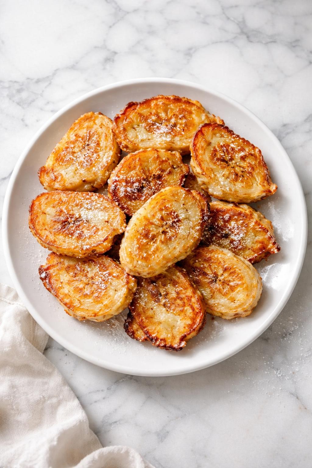 Image Prompt: Realistic top-down editorial food photography of vegan banana fritters on a white plate over a white marble countertop, golden pan-fried banana fritters dusted lightly with sugar, soft inside with crisp edges, bright natural lighting, cozy homemade dessert mood, no people, no hands, no text, no watermarks, no props with writing.