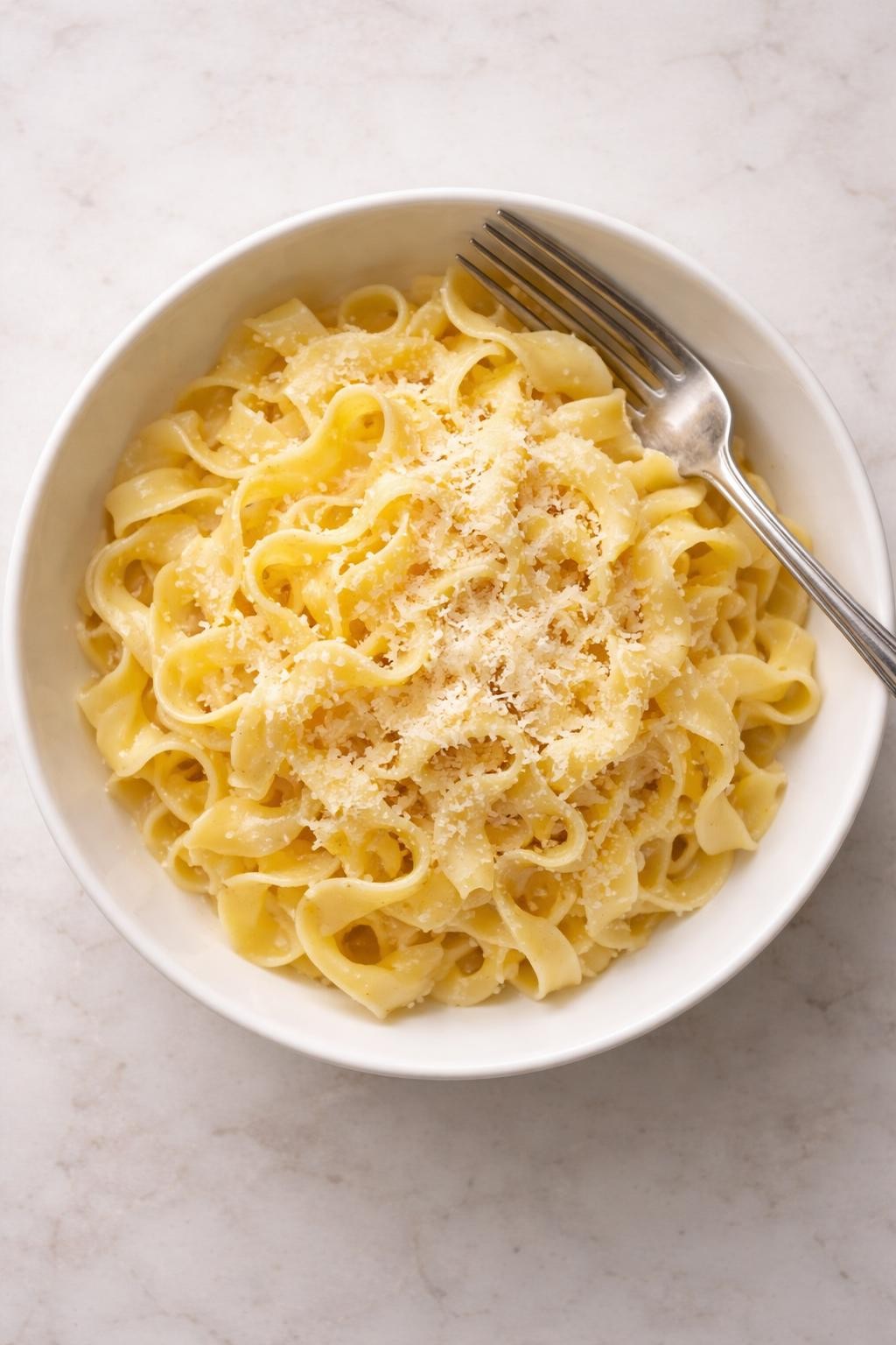"Top-down realistic food photography of a white bowl filled with plain egg noodles tossed in melted butter, with a light sprinkle of grated Parmesan cheese. No visible herbs or vegetables. A fork resting on the rim. Soft natural light, clean and simple comfort food, no people, no hands, no text, no watermarks."