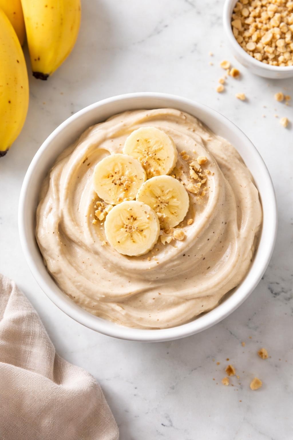 Image Prompt: Realistic top-down editorial food photography of vegan banana nice cream in a white bowl on a clean marble countertop, creamy frozen banana dessert scooped and topped with banana slices, smooth soft-serve texture, bright natural light, fresh simple dessert styling, no people, no hands, no text, no watermarks, no props with writing.