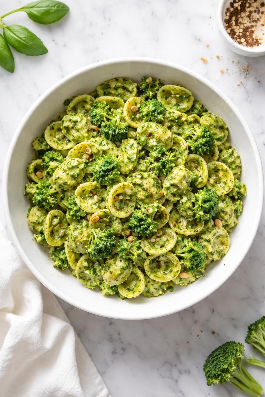 Image Prompt: Realistic top-down editorial food photography of vegan broccoli pesto pasta in a white bowl on a clean marble countertop, pasta coated in green broccoli pesto sauce with garlic and herbs, simple homemade dinner styling, bright natural light, no people, no hands, no text, no watermarks, no props with writing.