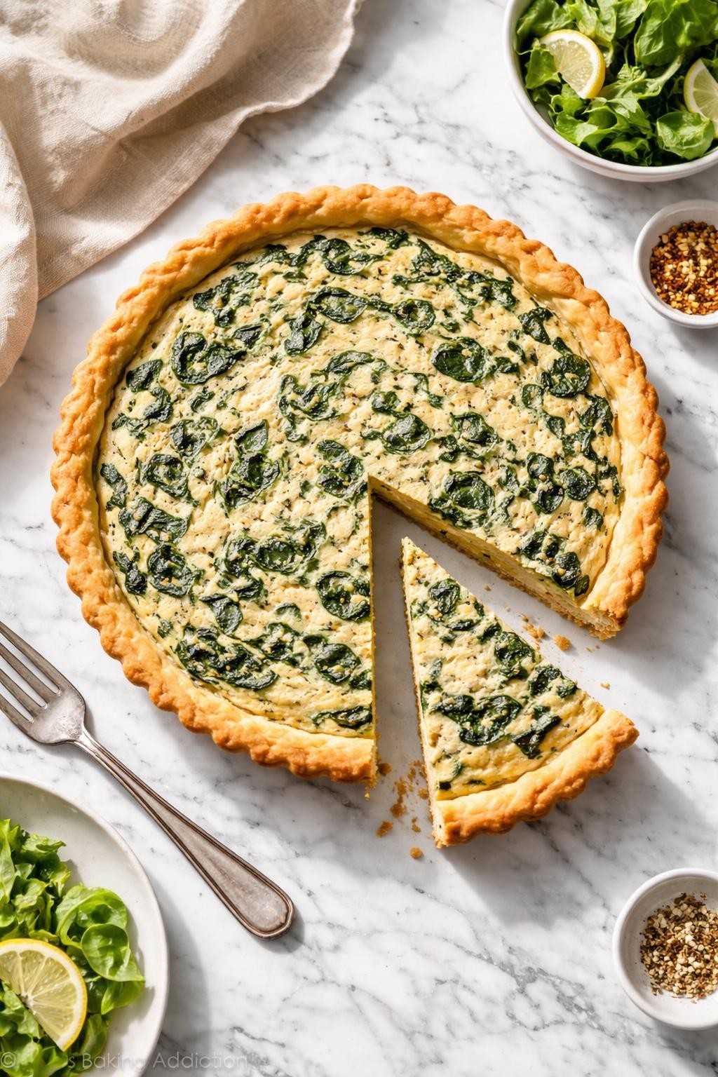 Image Prompt: Top-down realistic editorial food photography of a homemade vegan spinach quiche on a clean white and gray marble countertop, golden flaky crust filled with creamy tofu-based eggless filling and wilted spinach, one neat slice removed to show the soft savory center, warm cozy brunch mood, bright natural window lighting, soft shadows, clean composition, high detail, realistic food texture, no people, no hands, no text, no watermarks, no props with writing.