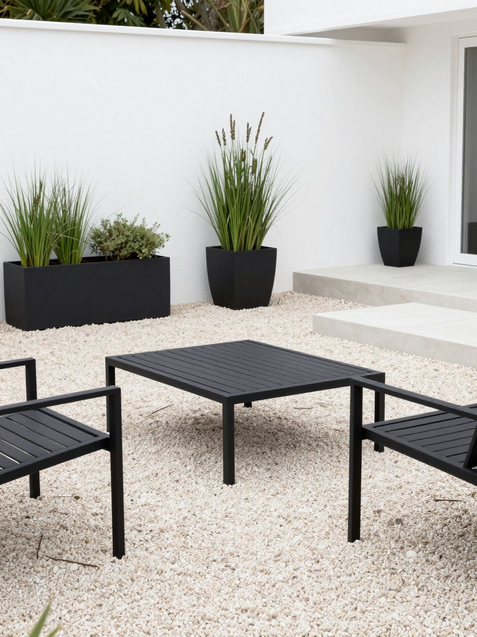 Image Prompt: A modern pea gravel patio with sleek black metal outdoor furniture, low rectangular coffee table, large black planters with ornamental grasses, pale pea gravel ground, white exterior wall, clean straight edging, stylish minimalist backyard design, no people, realistic DSLR photo, no text, no watermarks on images.