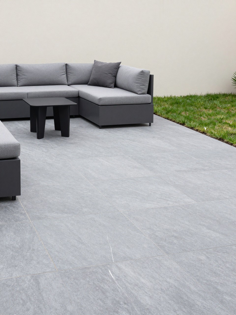 Image Prompt: A modern backyard patio with large smooth flagstone slabs in cool gray tones, simple low-profile outdoor sectional, black metal side table, sculptural planters, clean lawn edge, pale exterior wall, calm minimalist outdoor design, no people, realistic DSLR photo, no text, no watermarks on images.