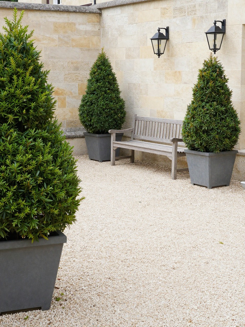 Image Prompt: A classic European patio with light pea gravel flooring, clipped boxwood shrubs in large planters, simple wooden bench, black iron lanterns, pale stone wall, neat formal garden mood, bright daylight, no people, realistic DSLR garden photo, no text, no watermarks on images.