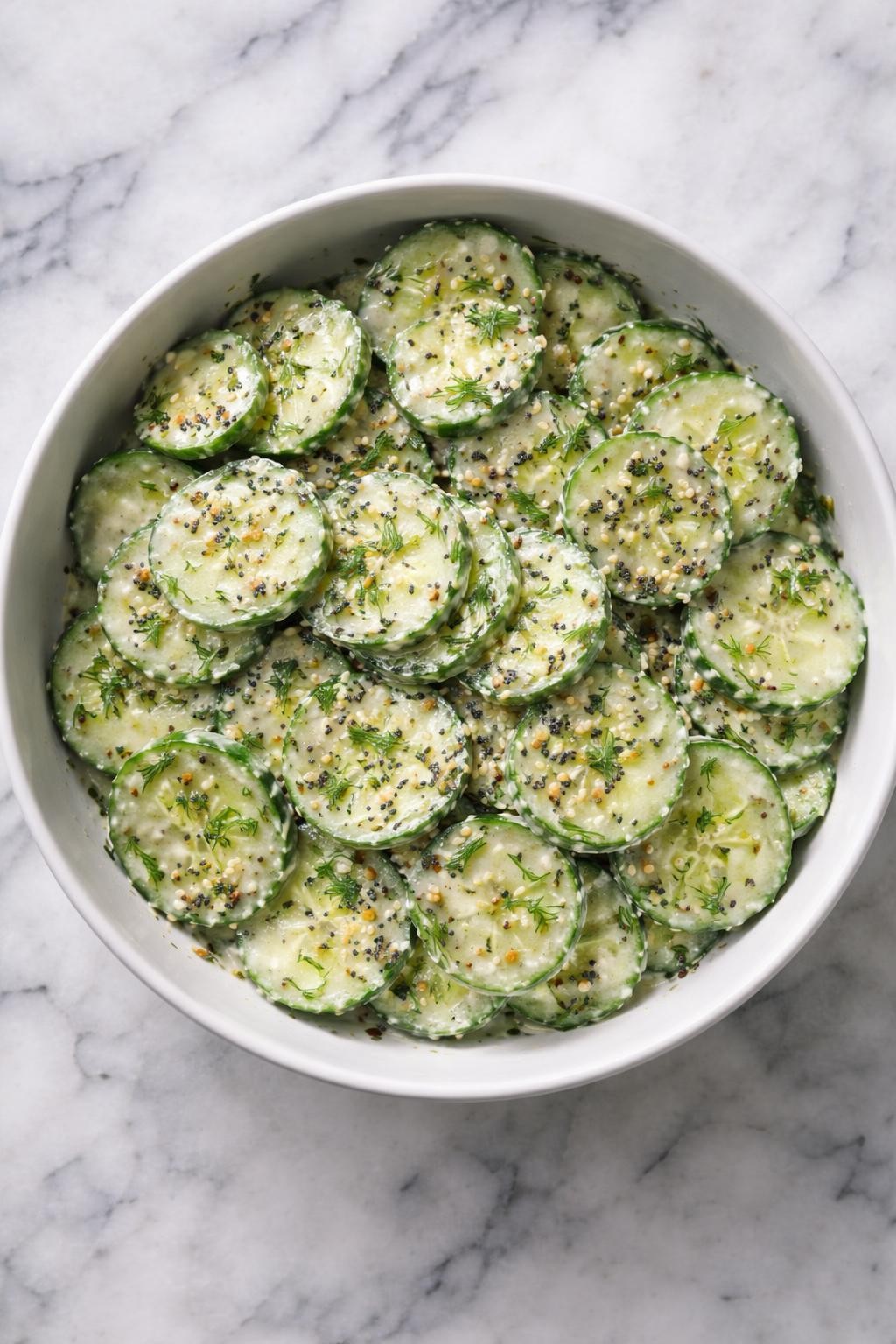 Image Prompt: Realistic top-down editorial food photography of everything bagel cucumber salad in a white bowl on a clean white and gray marble countertop, cucumber slices tossed in a creamy dressing and topped with everything bagel seasoning, chilled fresh salad mood, bright natural daylight, no people, no hands, no text, no watermarks, no props with writing.
