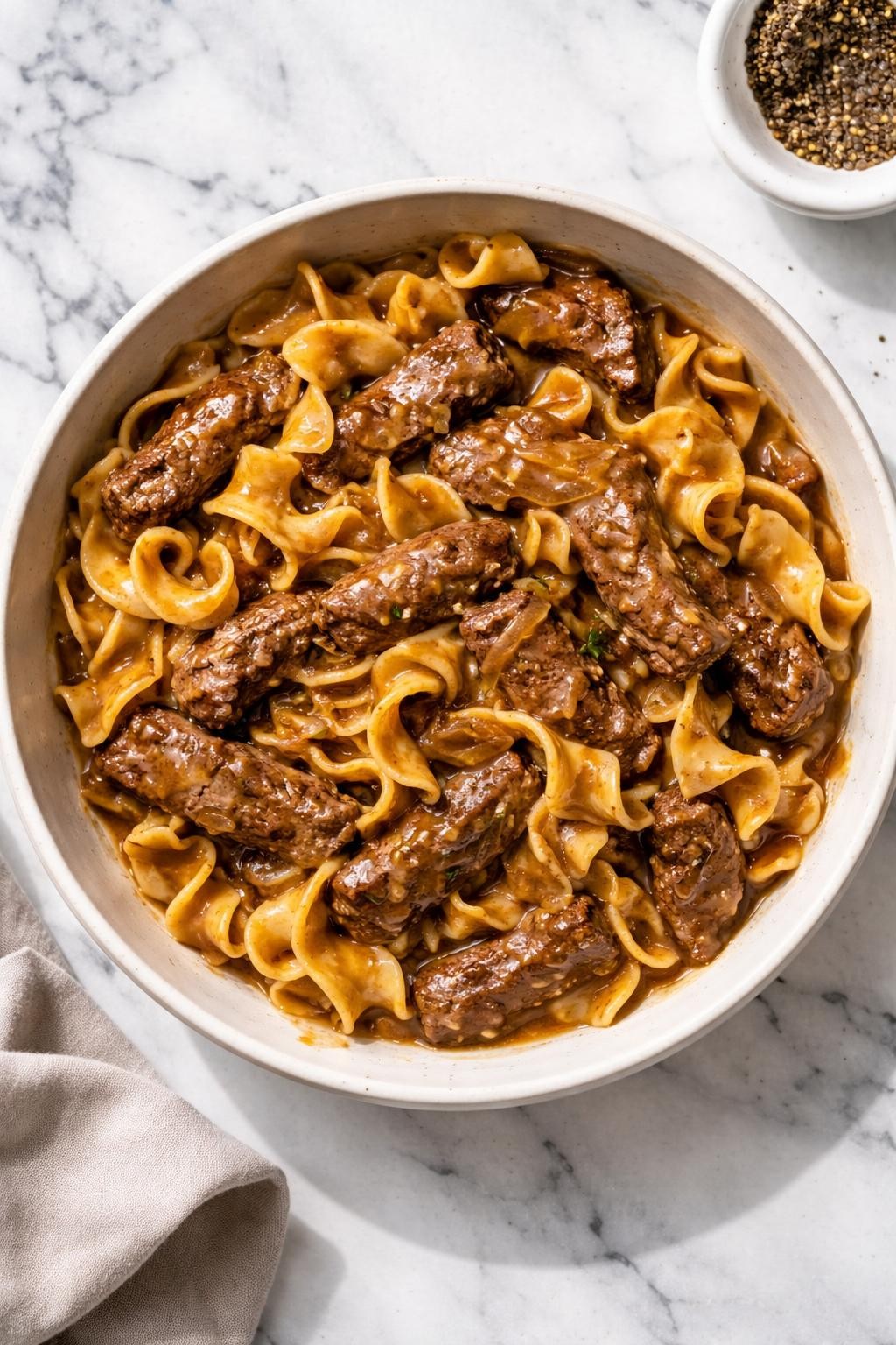 Image Prompt: Realistic top-down editorial food photography of onion soup mix beef and noodles on a white marble countertop, tender beef strips and egg noodles coated in a rich savory onion sauce, cozy homemade comfort food styling, bright natural daylight, soft shadows, clean editorial composition, high detail, realistic texture, no people, no hands, no text, no watermarks, no props with writing.