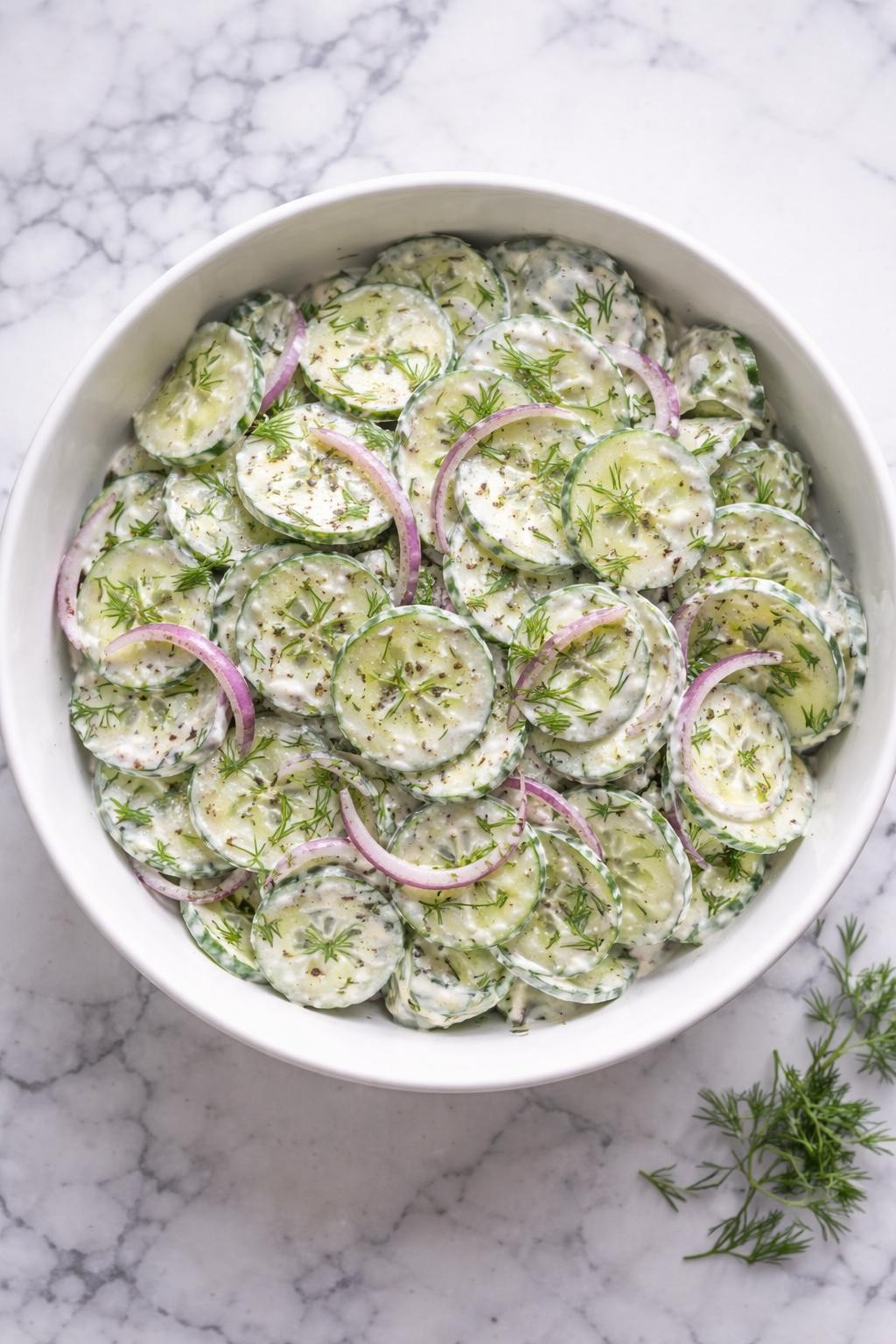 Image Prompt: Realistic top-down editorial food photography of creamy TikTok cucumber salad in a white serving bowl on a clean white and gray marble countertop, thinly sliced cucumbers coated in a creamy dressing with sour cream, fresh dill, black pepper, and red onion, chilled fresh salad mood, bright natural daylight, clean composition, high detail, realistic food texture, no people, no hands, no text, no watermarks, no props with writing.