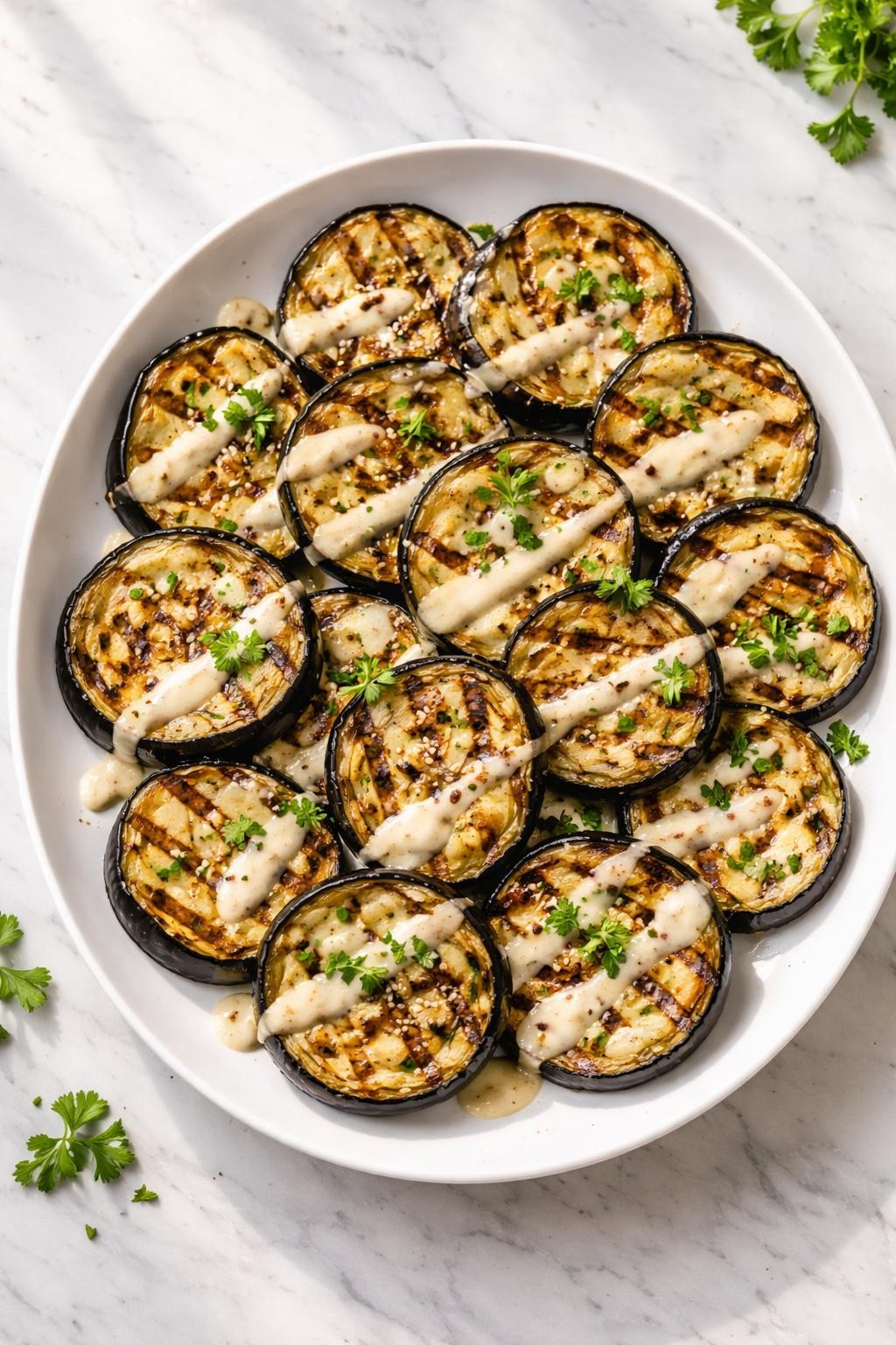 Image Prompt: Realistic top-down editorial food photography of grilled eggplant slices drizzled with lemon tahini sauce on a white serving plate on a clean marble countertop, charred tender eggplant rounds with creamy pale sauce, parsley, and sesame seeds, bright natural window lighting, soft shadows, clean composition, high detail, realistic food texture, no people, no hands, no text, no watermarks, no props with writing.
