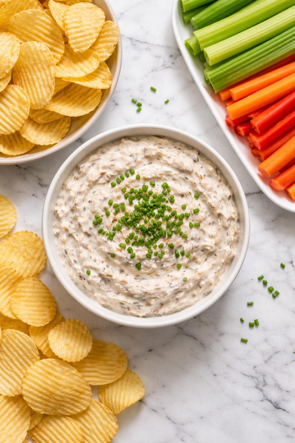 Image Prompt: Realistic top-down editorial food photography of a bowl of homemade French onion dip on a clean white and gray marble countertop, creamy dip topped with chopped chives, surrounded by potato chips and raw vegetable sticks, simple party snack styling, bright natural window lighting, soft shadows, clean editorial composition, high detail, realistic texture, no people, no hands, no text, no watermarks, no props with writing.