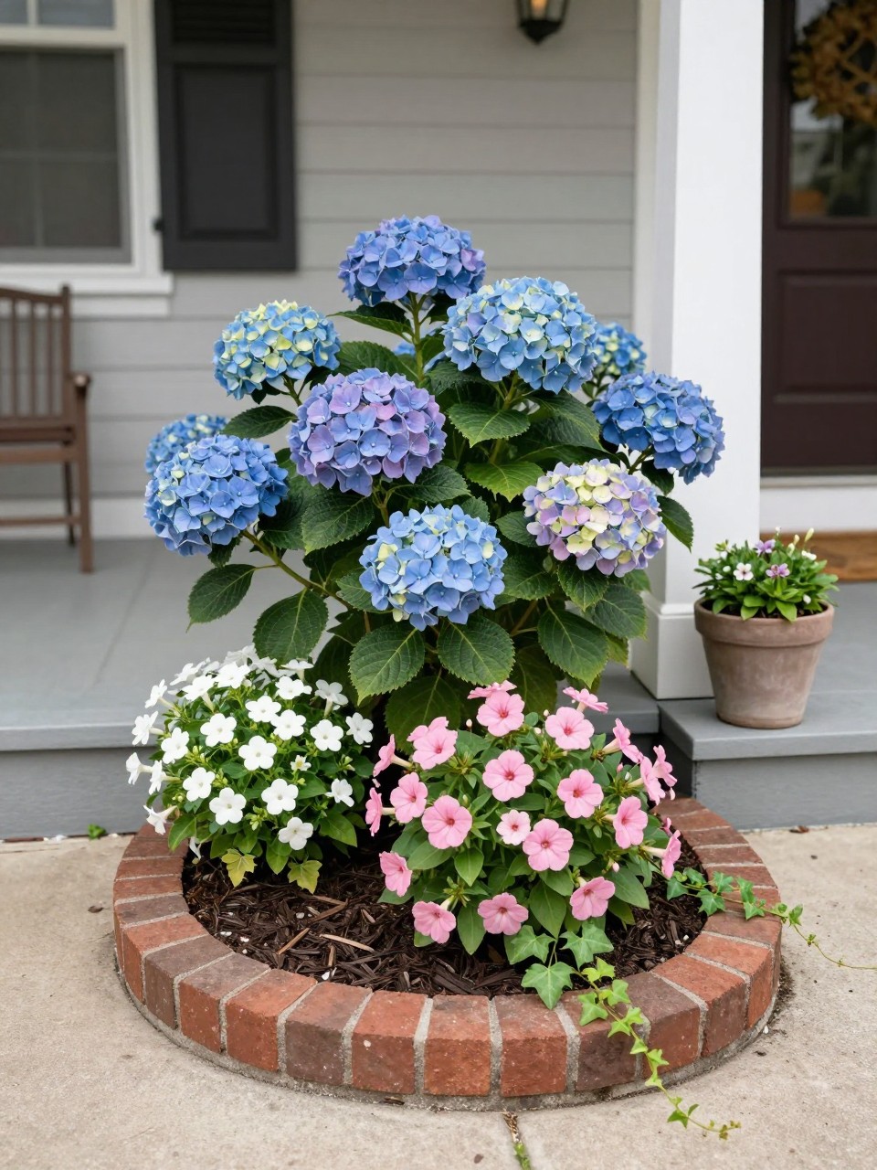 A small flower bed beside a front porch, filled with hydrangeas, pink impatiens, white begonias, and trailing ivy, dark mulch, curved brick edging, cozy porch steps with simple planters nearby, soft daylight, realistic welcoming home exterior, no people, no text, no watermark.