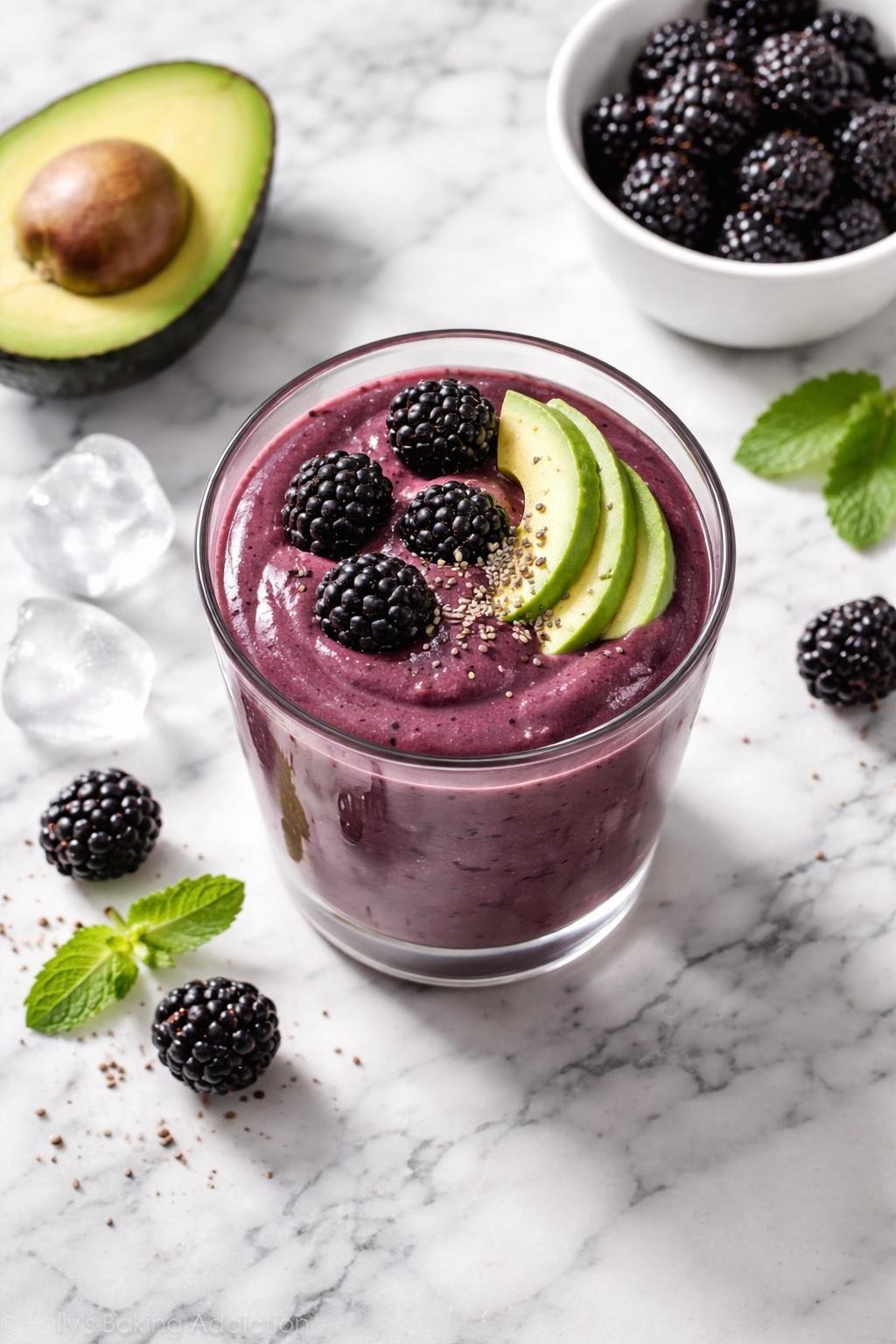 Realistic top-down editorial food photography of a blackberry avocado low carb smoothie served in a clear glass on a clean white and gray marble countertop. The smoothie is deep purple, thick, creamy, and smooth, styled with blackberries, avocado slices, chia seeds, and a few ice cubes nearby. Bright natural daylight, clean berry smoothie styling, realistic creamy texture, high detail, no people, no hands, no text, no watermarks, no props with writing.