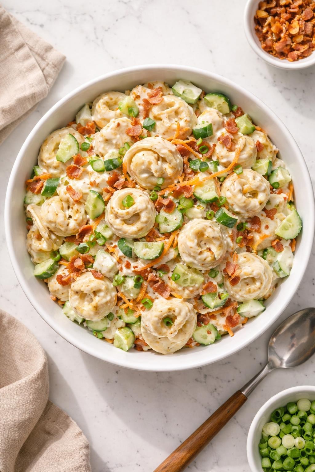 Image Prompt: Realistic top-down editorial food photography of creamy tortellini ranch pasta salad in a simple serving bowl on a white marble countertop, cheese tortellini coated in creamy ranch dressing with chopped cucumber, shredded carrots, bacon bits, and green onions, cozy fresh homemade mood, bright natural light, soft shadows, clean composition, no people, no hands, no text, no watermarks, no props with writing.