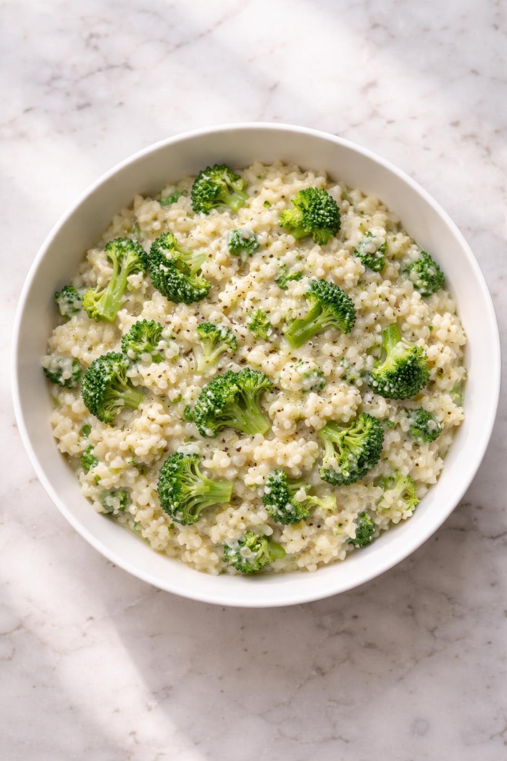 Image Prompt: Top-down realistic editorial food photography of vegan broccoli risotto in a white bowl on a clean marble countertop, creamy arborio rice with chopped tender broccoli folded through, glossy dairy-free texture, simple homemade dinner styling, natural daylight, soft shadows, high detail, no people, no hands, no text, no watermarks, no props with writing.