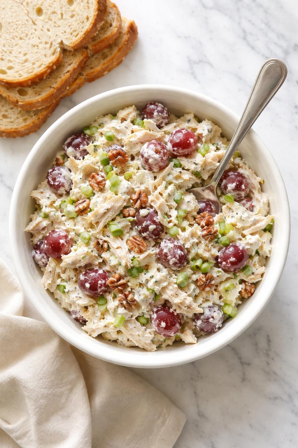 "Overhead realistic photo of a white bowl filled with chicken salad made with Greek yogurt, shredded chicken, red grapes, and chopped pecans. A spoon resting on the rim. A stack of bread slices next to the bowl. Bright natural light, creamy make-ahead lunch, no people, no hands, no text, no watermarks."