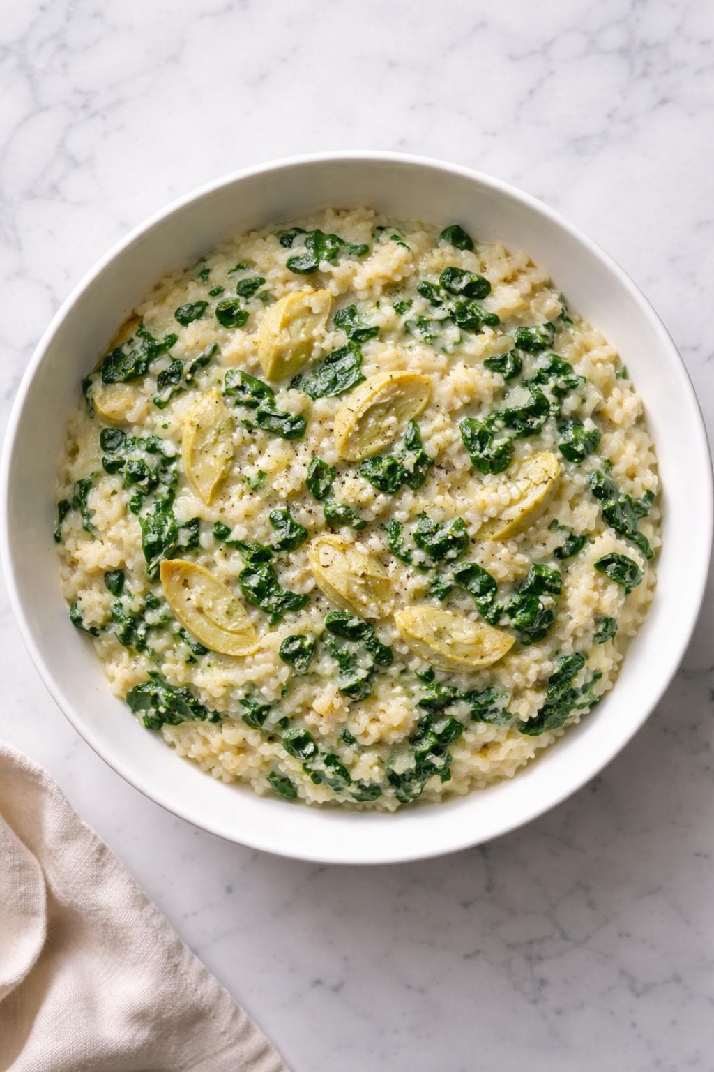 Image Prompt: Top-down realistic editorial food photography of vegan spinach artichoke risotto in a white bowl on a clean marble countertop, creamy arborio rice with chopped spinach and artichoke hearts folded through, rich glossy texture, homemade comfort dinner styling, bright natural light, soft shadows, high detail, no people, no hands, no text, no watermarks, no props with writing.