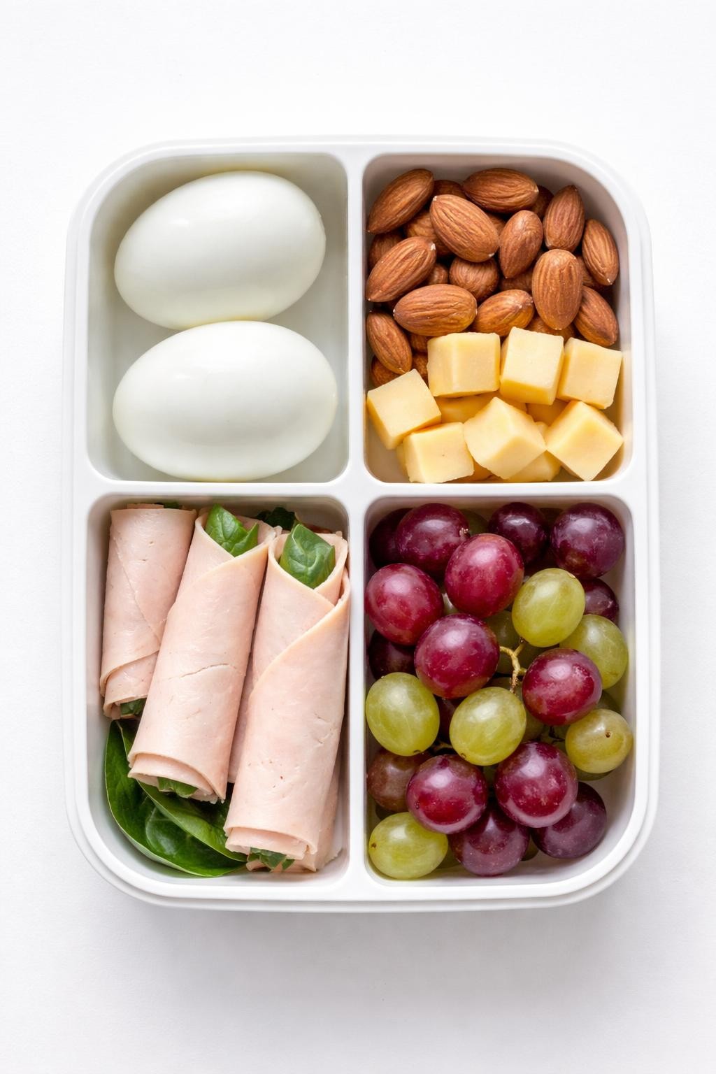"Top-down realistic food photography of a bento-style lunch box with compartments: two peeled hard-boiled eggs, a handful of almonds, a few slices of turkey roll-ups, cheese cubes, and grapes. Clean white background, bright daylight, portable high-protein snack prep, no people, no hands, no text, no watermarks."