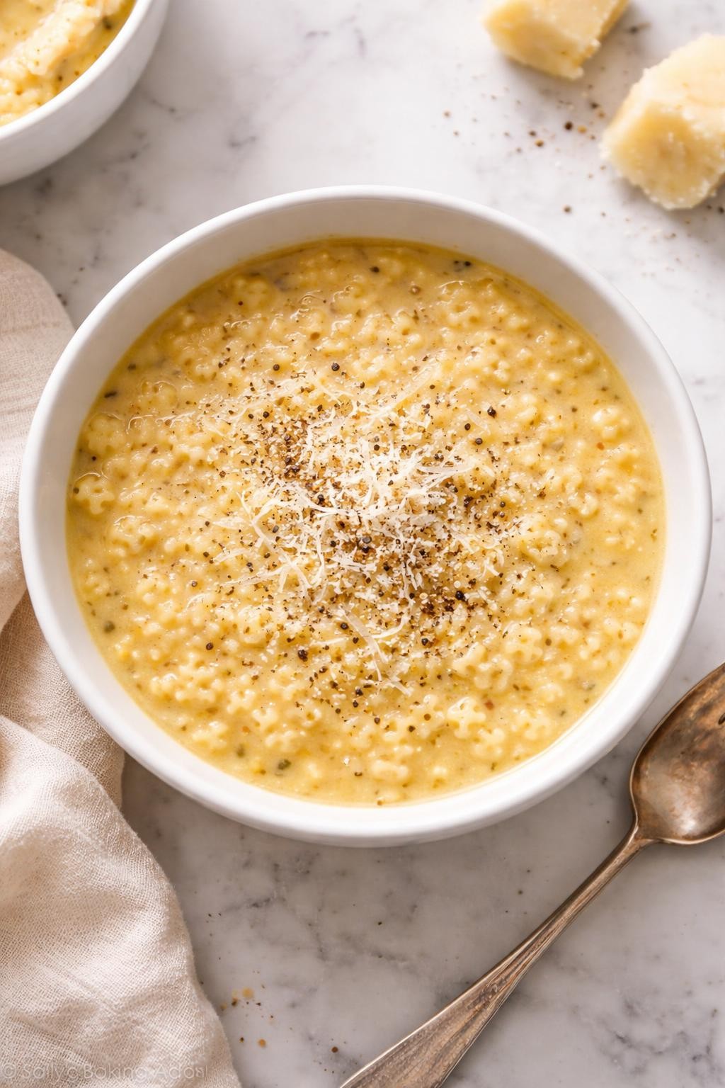 Image Prompt: Realistic top-down editorial food photography of parmesan pastina soup in a white bowl on a white marble countertop, creamy golden broth filled with tiny pastina pasta and topped with extra grated parmesan and black pepper, cozy homemade soup styling, bright natural light, no people, no hands, no text, no watermarks, no props with writing.