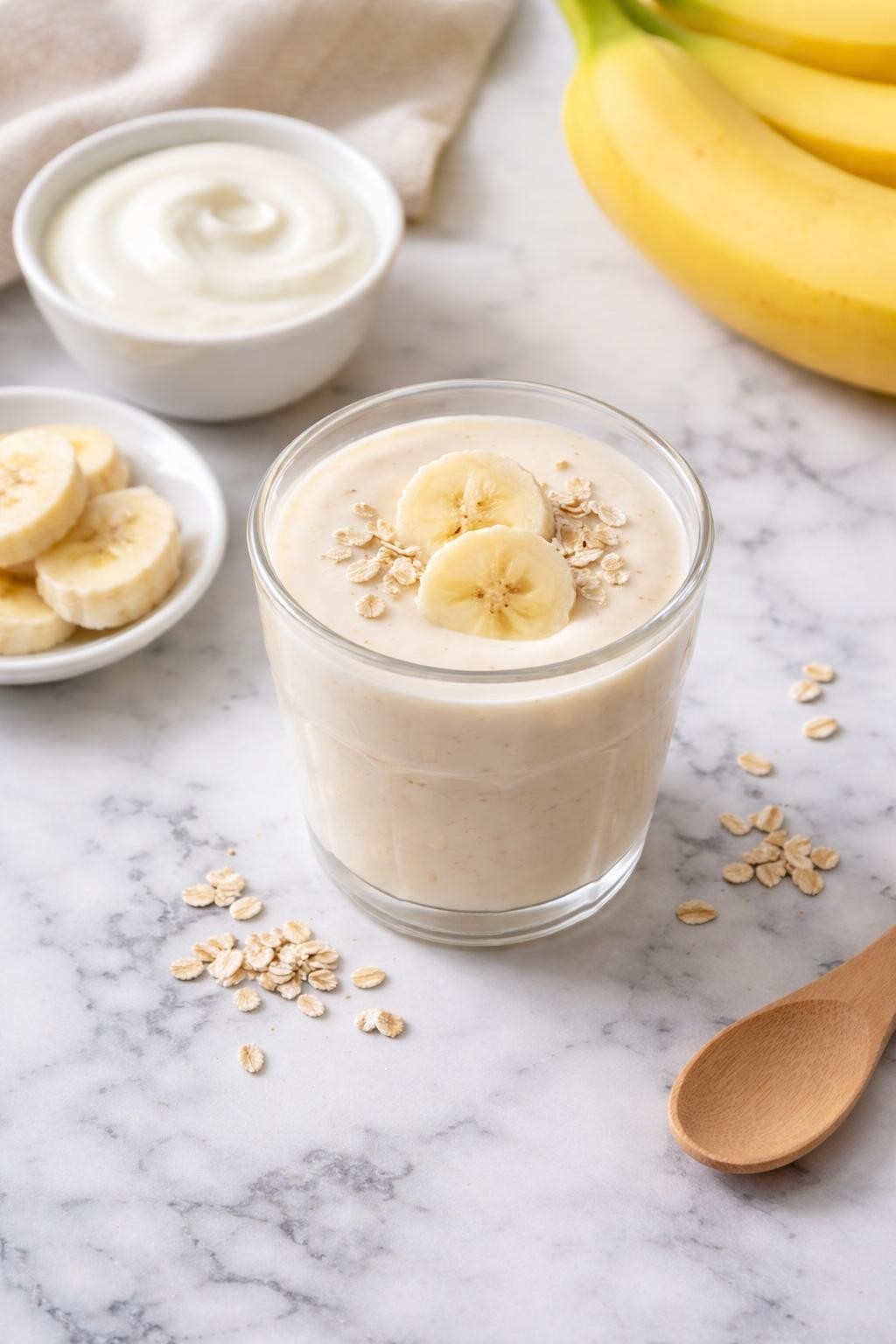 Realistic top-down editorial food photography of a small banana yogurt toddler smoothie served in a short clear toddler-safe cup on a clean white and gray marble countertop. The smoothie is pale cream, smooth, thick, and creamy, with banana slices, a small plain bowl of yogurt, and a few soft oat flakes styled neatly nearby. Bright natural window lighting, soft shadows, clean family snack styling, high detail, realistic smoothie texture, no people, no hands, no text, no watermarks, no props with writing.