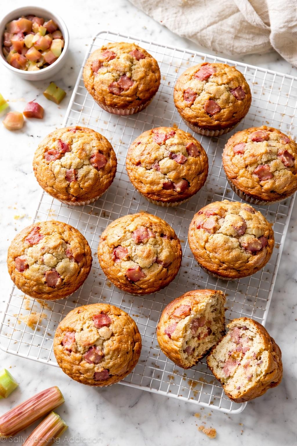 Image Prompt: Top-down realistic food photography of freshly baked vegan rhubarb muffins arranged on a white cooling rack over a light marble countertop, golden muffin tops with visible pink rhubarb pieces, one muffin broken open to show the soft fluffy crumb, bright natural daylight, cozy homemade bakery mood, clean editorial styling, no people, no hands, no text, no watermarks, no props with writing.