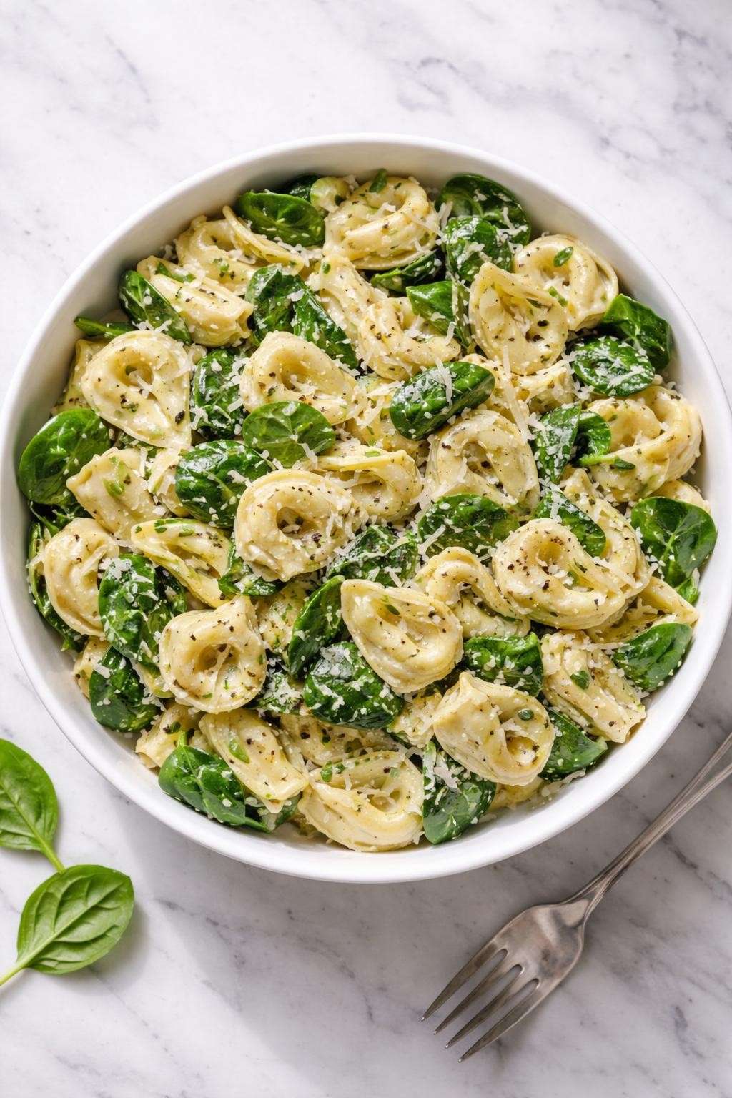 Image Prompt: Realistic top-down editorial food photography of spinach and artichoke tortellini pasta salad in a large white bowl on a clean marble countertop, cheese tortellini tossed with chopped artichoke hearts, baby spinach, parmesan, and creamy dressing, bright natural window lighting, soft shadows, clean composition, no people, no hands, no text, no watermarks, no props with writing.